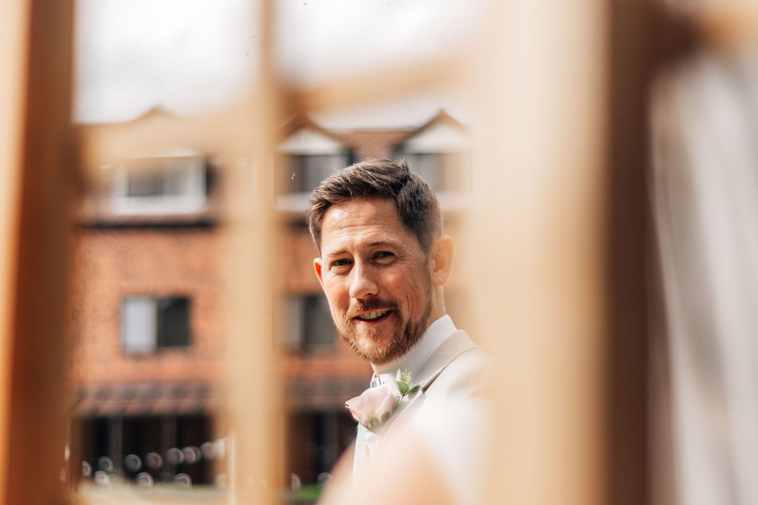 Groom smiling warmly in white suit with floral boutonniere, photographed through wooden frame at outdoor wedding venue.