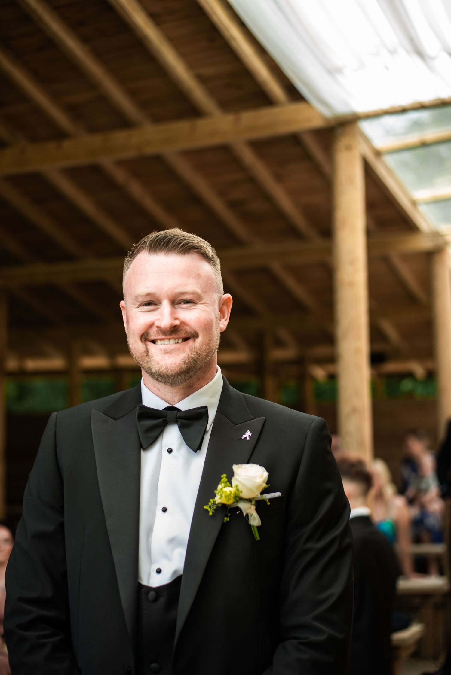 Smiling groom in black tuxedo with bow tie and white boutonniere stands at outdoor wedding venue.