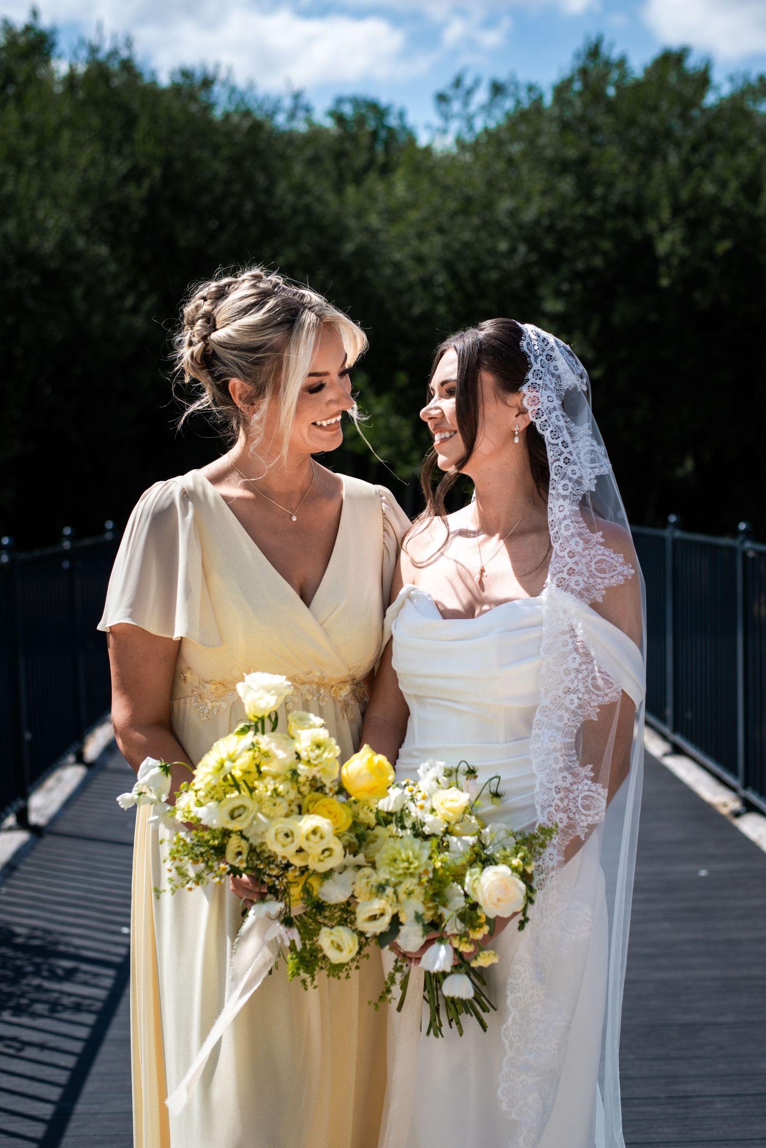 Bride in white gown and lace veil smiling with bridesmaid in yellow dress holding bouquets on bridge.