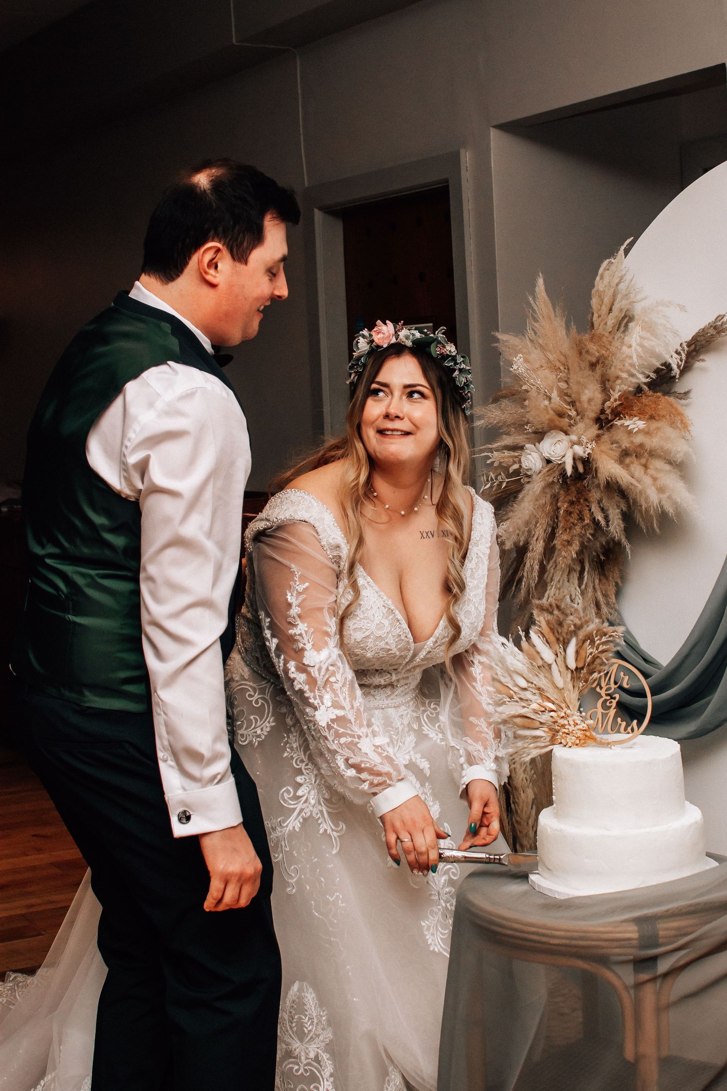 Bride and groom cutting white wedding cake, bride wearing lace gown and floral crown, pampas grass decor in background.
