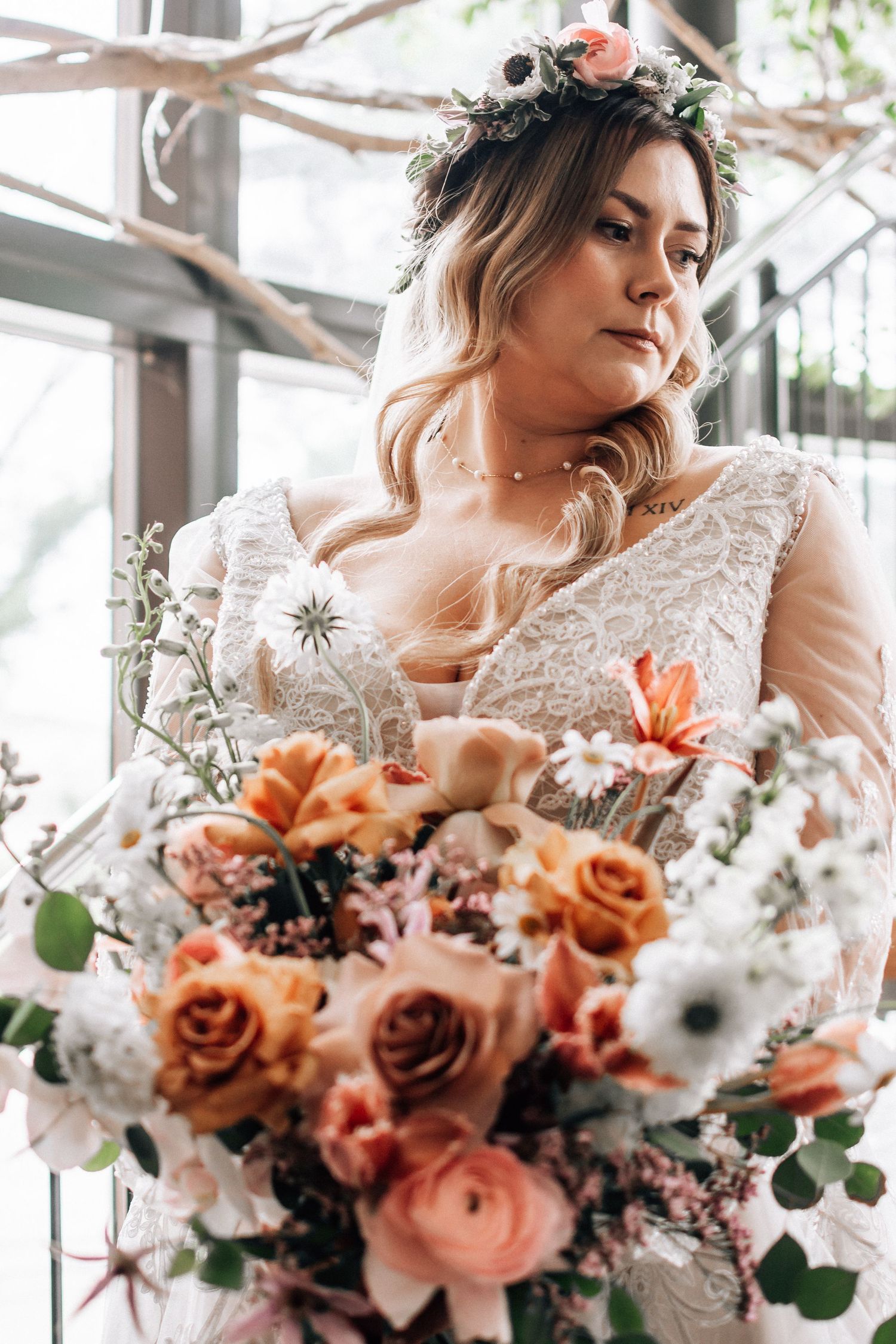 Bride in lace gown holds lush peach and white floral bouquet, wearing a flower crown indoors.