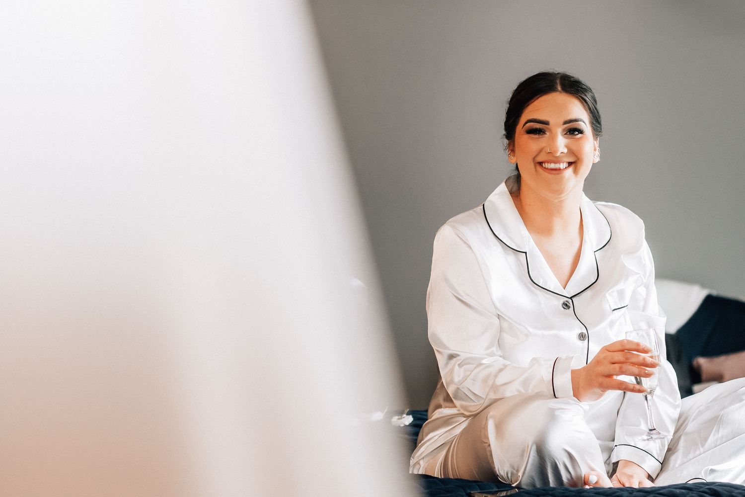 Smiling bride in white satin pajamas sitting on a bed, holding a glass during bridal preparations.
