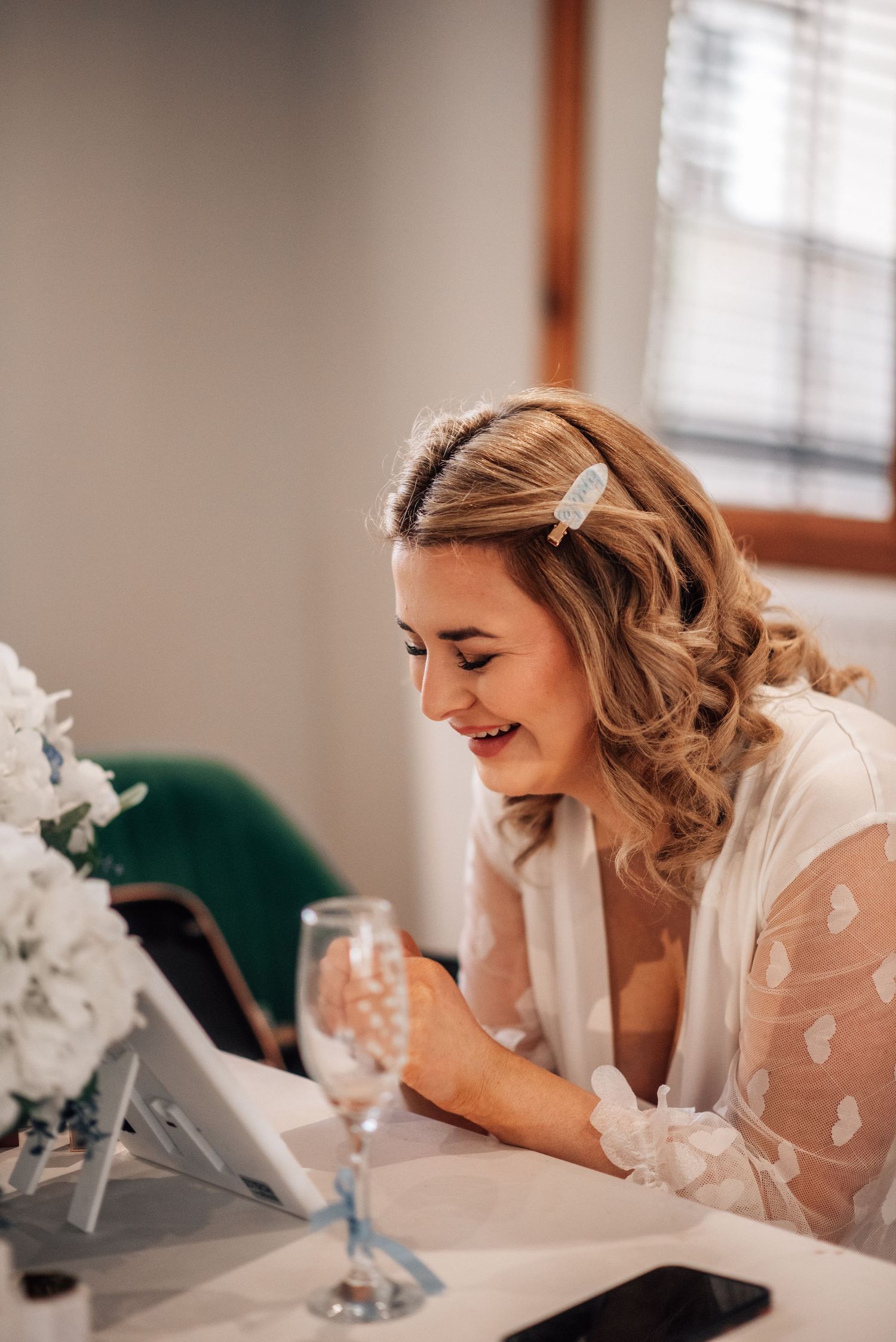 Smiling bride in white robe with hair bow accessory looking at mirror on wedding morning preparation.