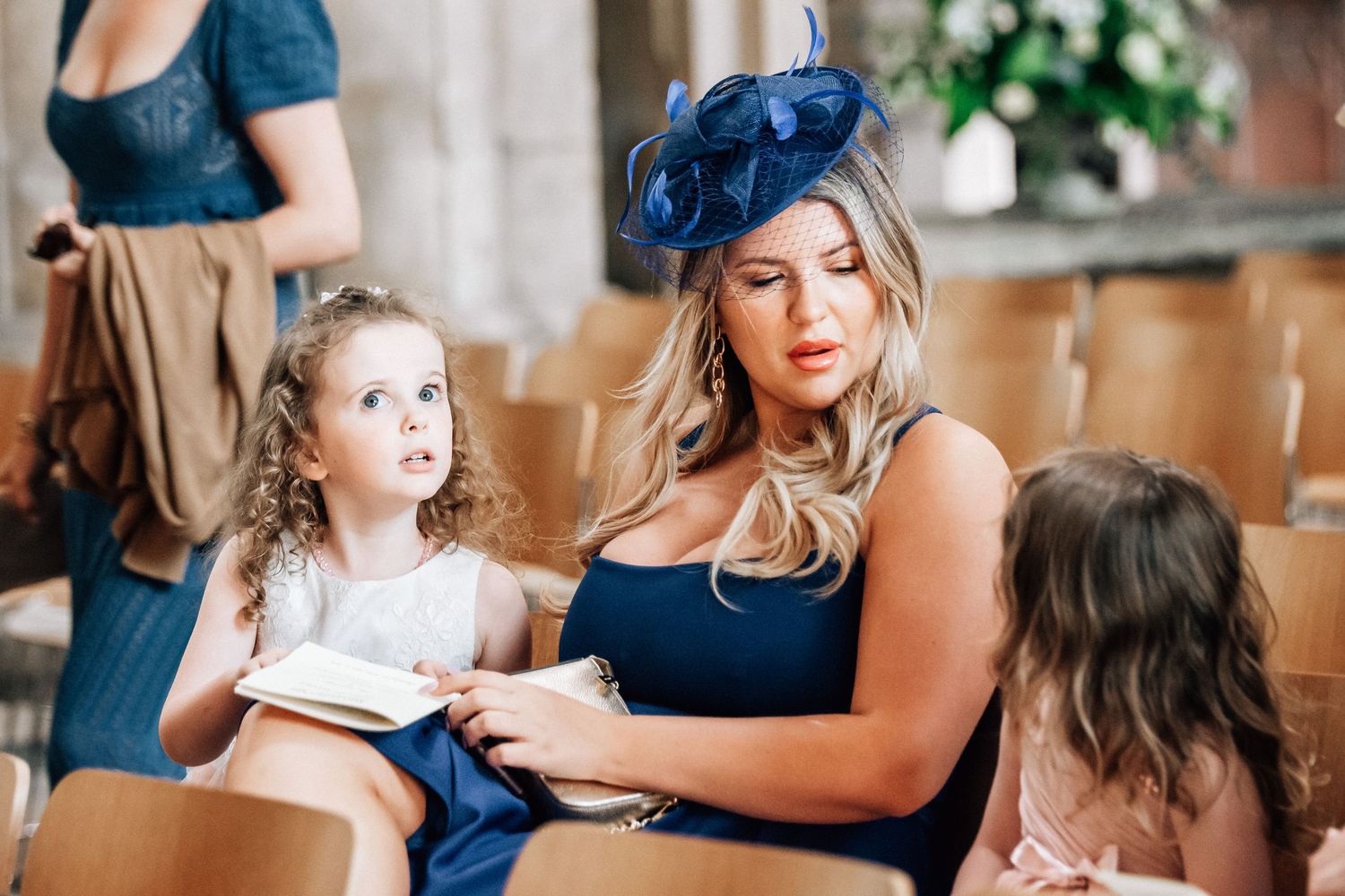 Woman in navy dress and fascinator sits with young girl in white dress at a wedding ceremony.