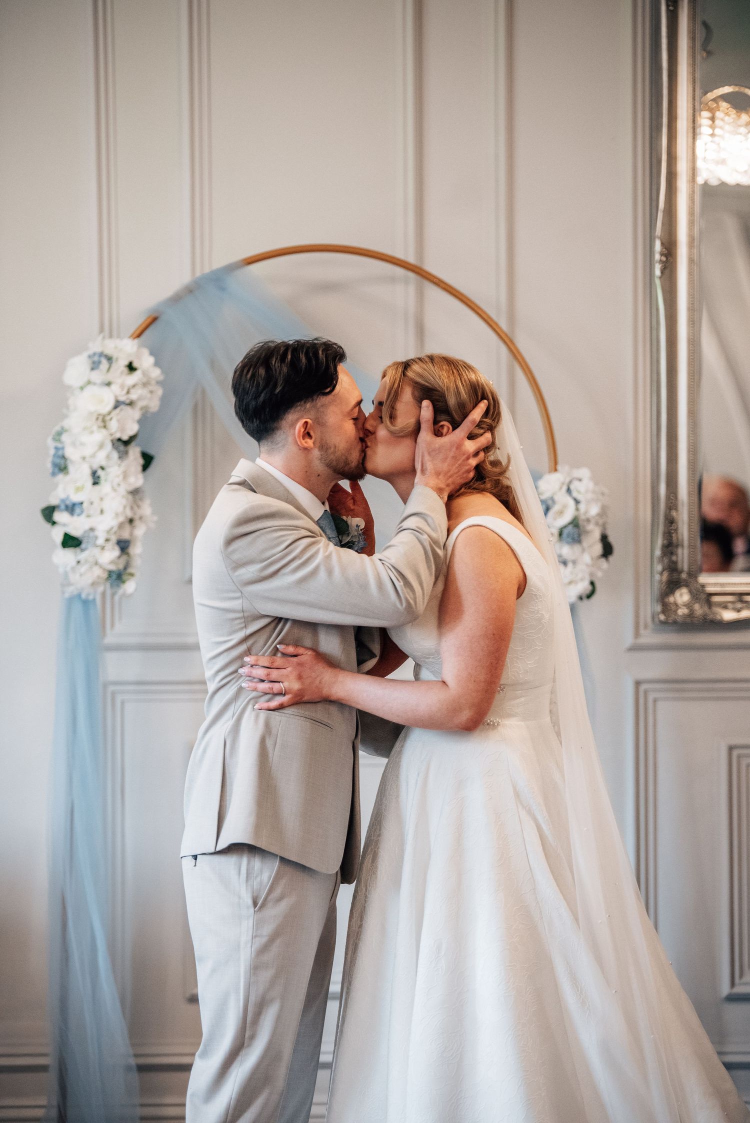 Bride and groom share first kiss at altar with circular floral arch and white roses in elegant indoor venue.