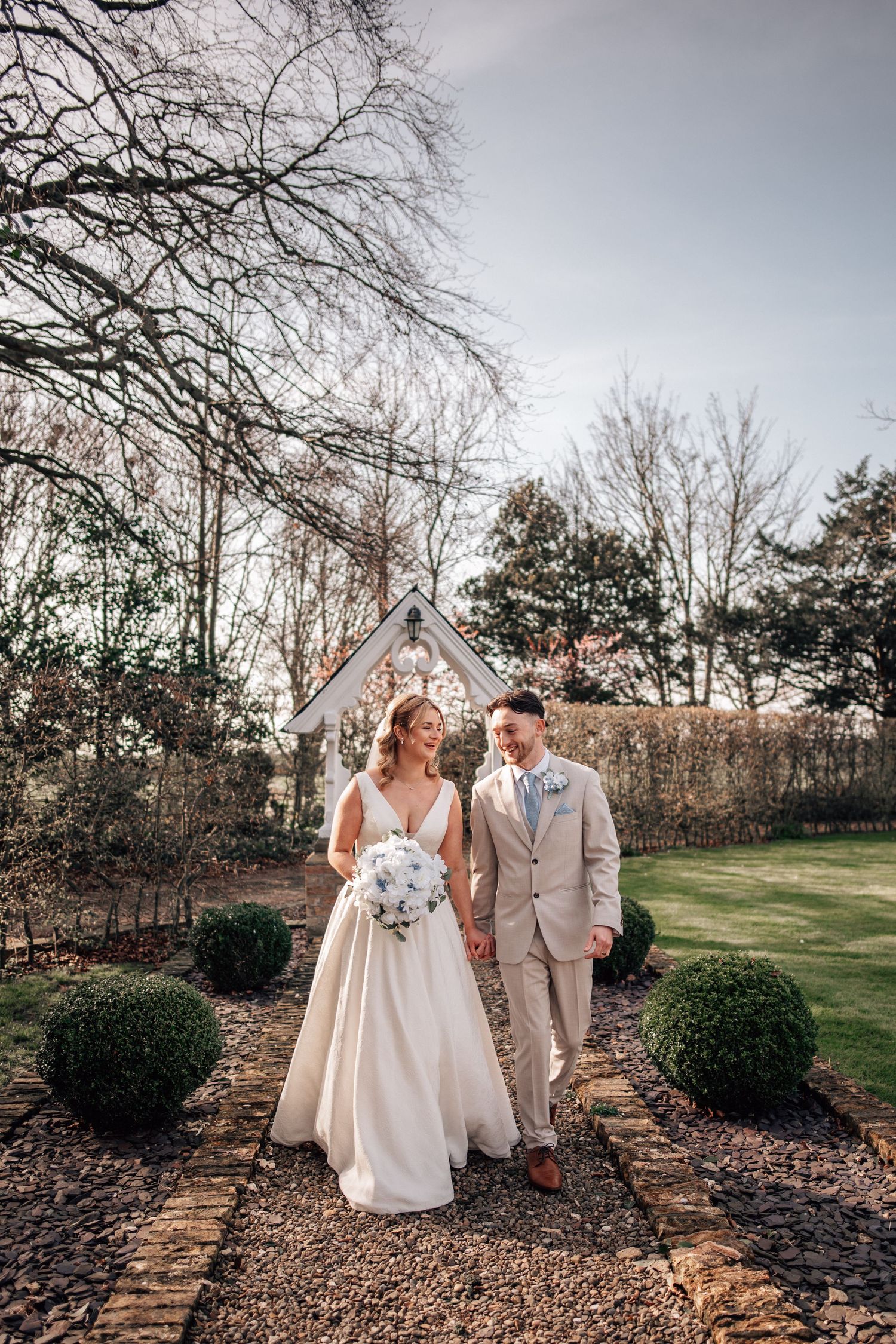 Bride and groom walk hand-in-hand through a garden path with a white chapel backdrop on their wedding day.