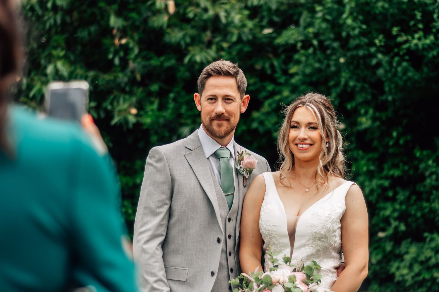 A smiling bride and groom pose together outdoors in front of lush greenery while a guest photographs them on a smartphone.