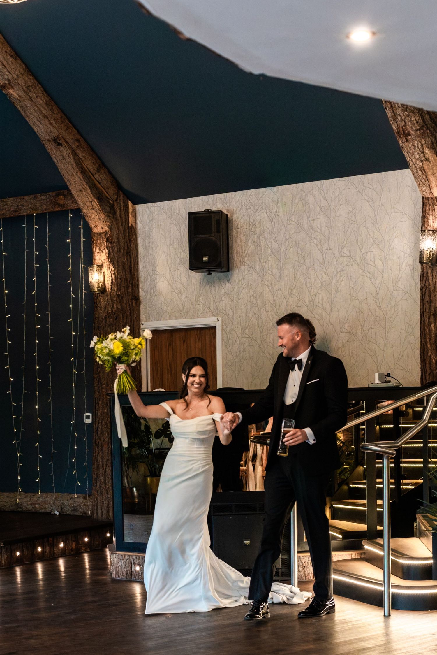 Joyful bride in white gown raising bouquet while groom in tuxedo holds her hand at wedding venue.