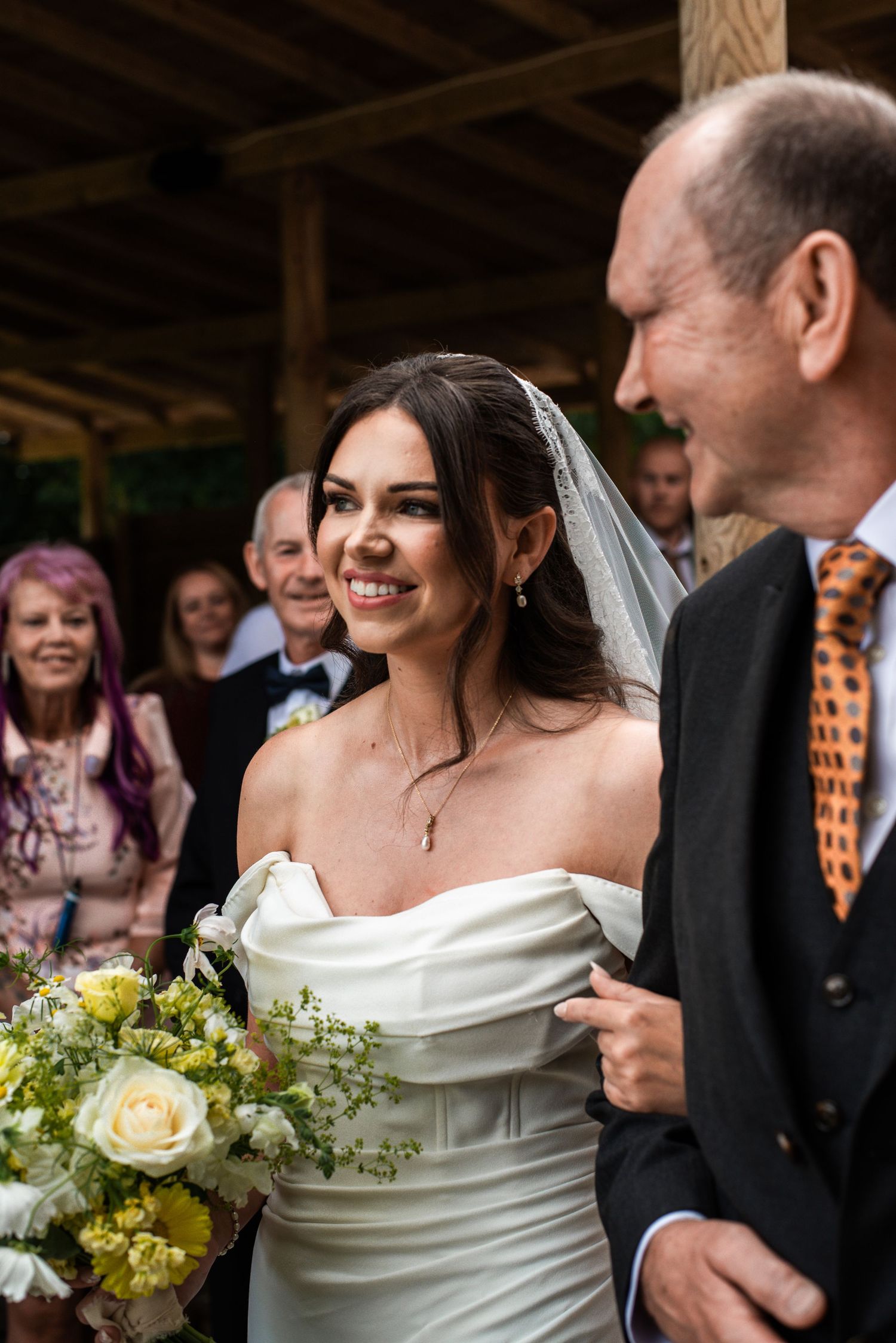 A smiling bride in an off-shoulder white gown holds a bouquet while standing with guests at her wedding.