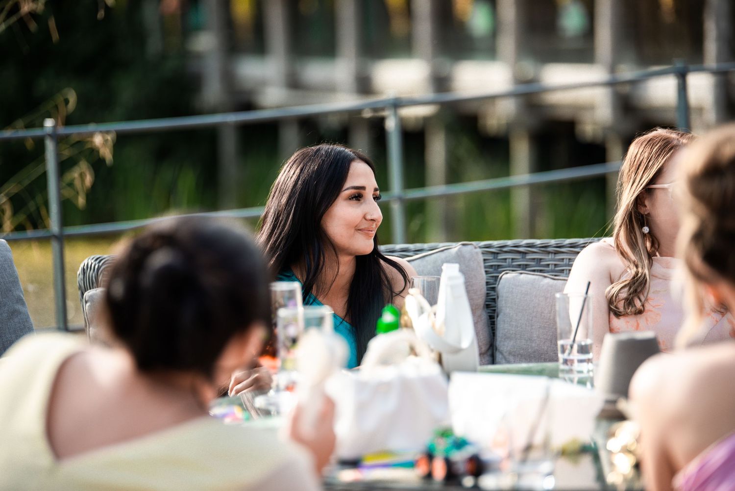 Woman with dark hair smiling at an outdoor wedding reception dinner table with guests and greenery in background.
