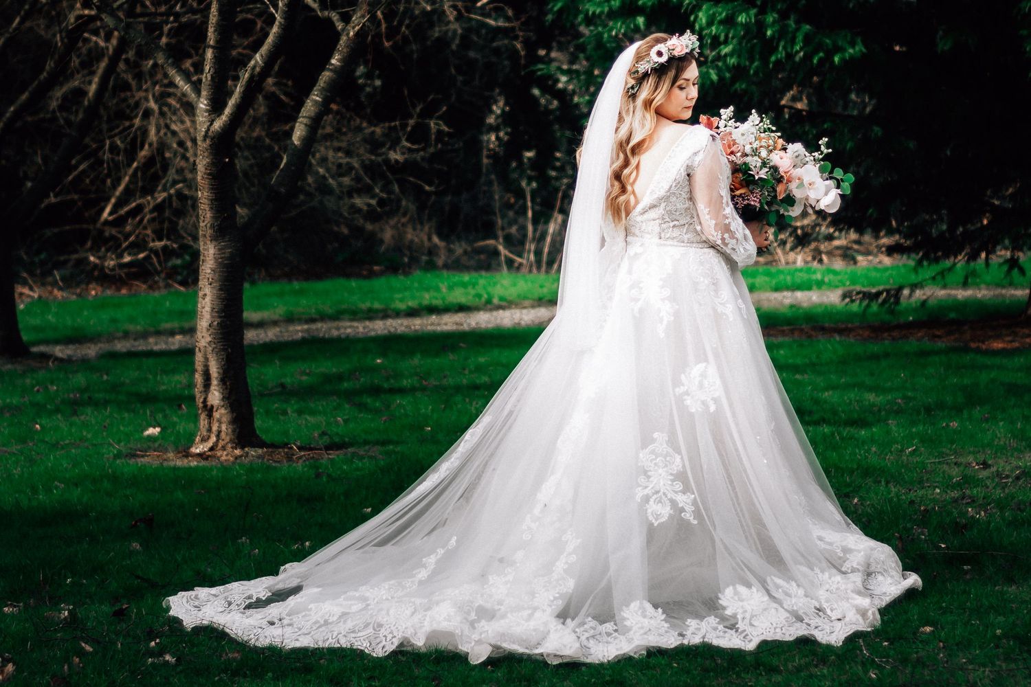Bride in white lace wedding gown with long train and veil holds bouquet outdoors on green grass.