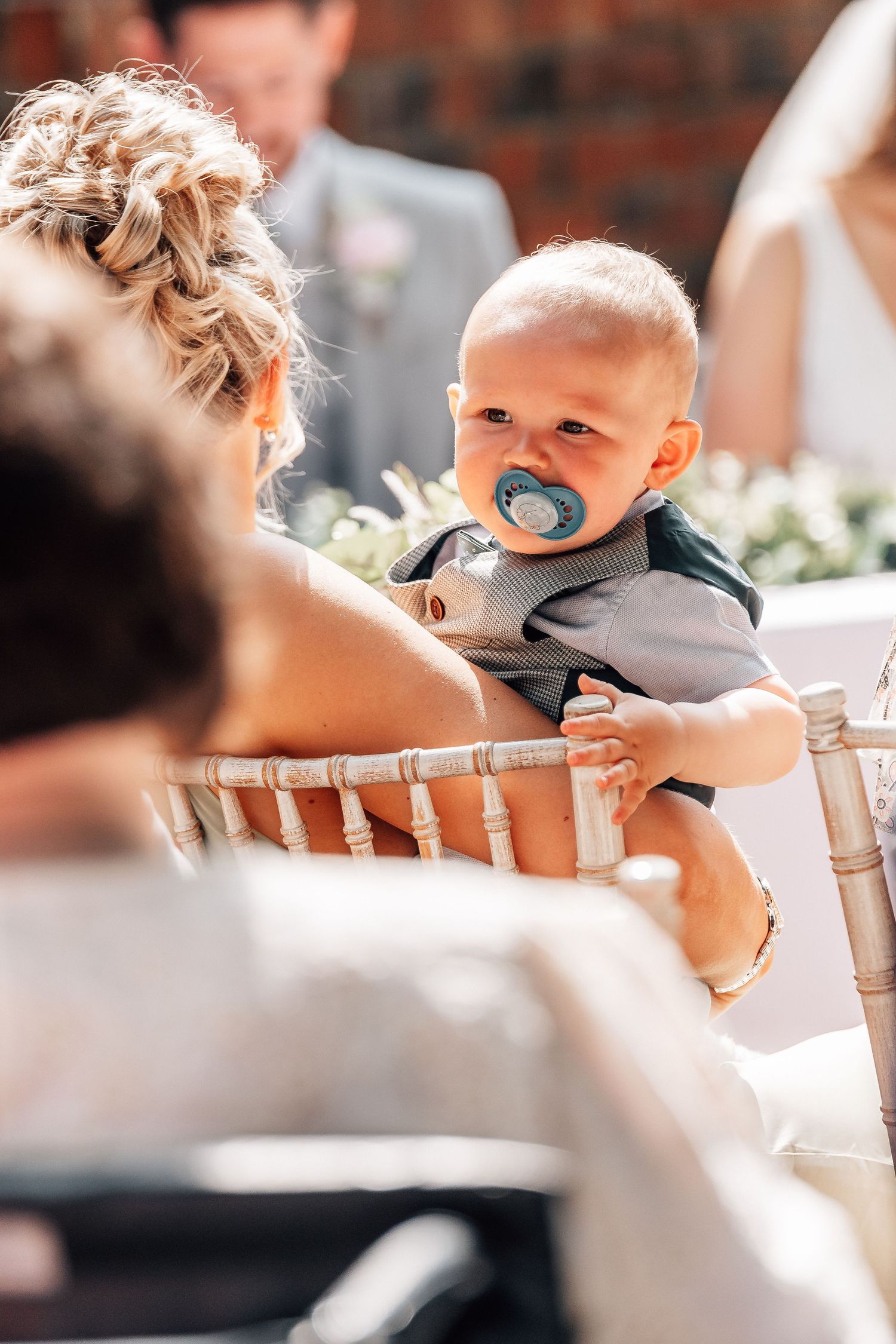 Baby with pacifier sitting on lap at outdoor wedding ceremony.