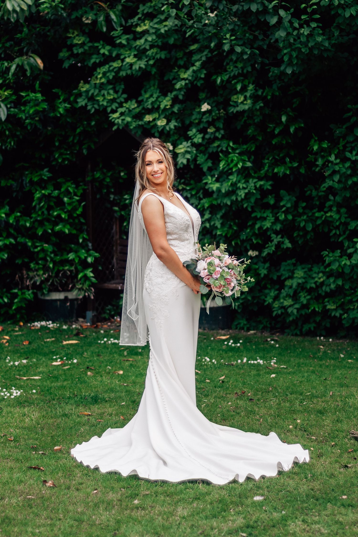 Radiant bride in a fitted white lace gown holding a pink bouquet in a lush garden setting.
