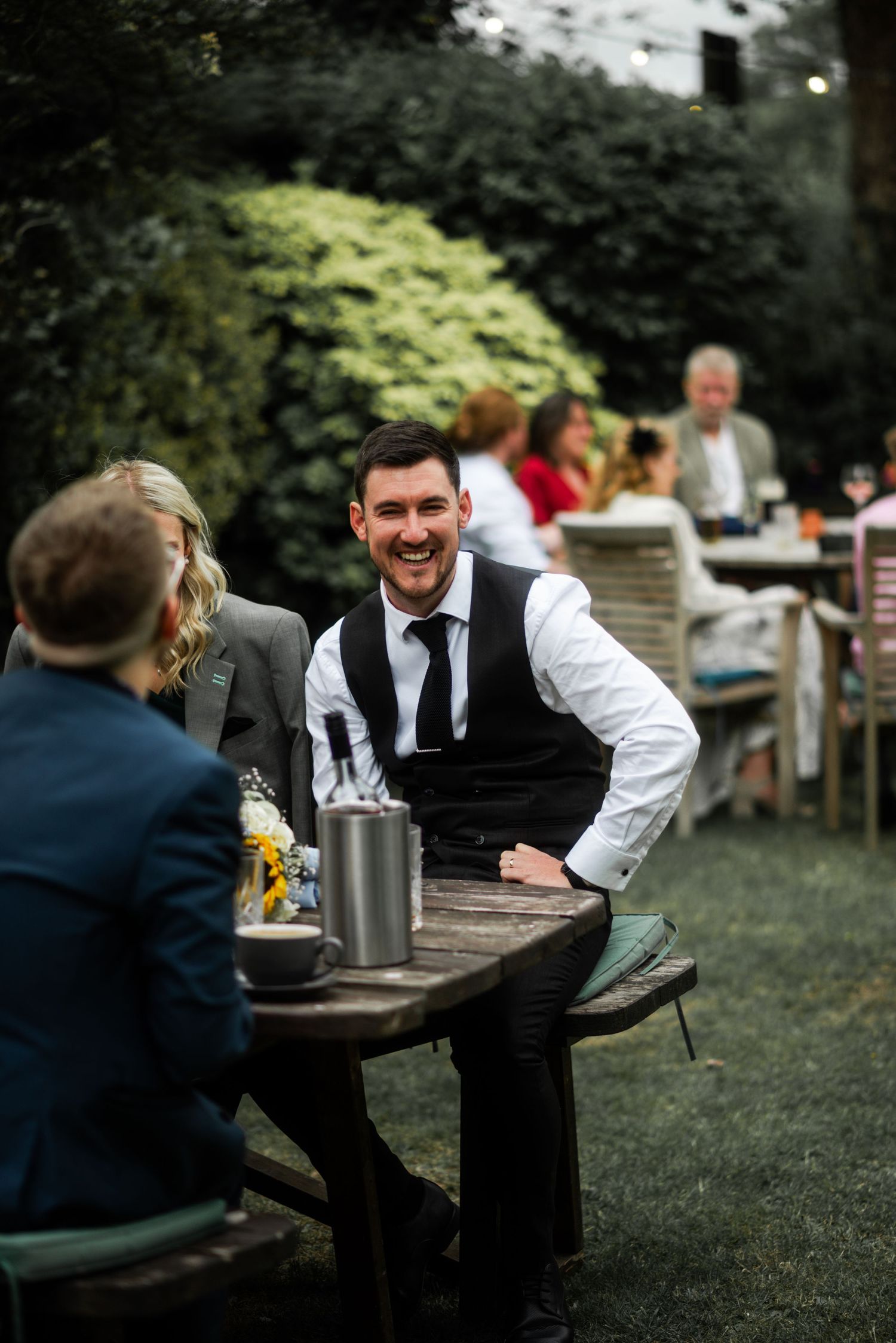 Smiling groom in vest and tie laughs with guests at outdoor wedding reception garden tables.