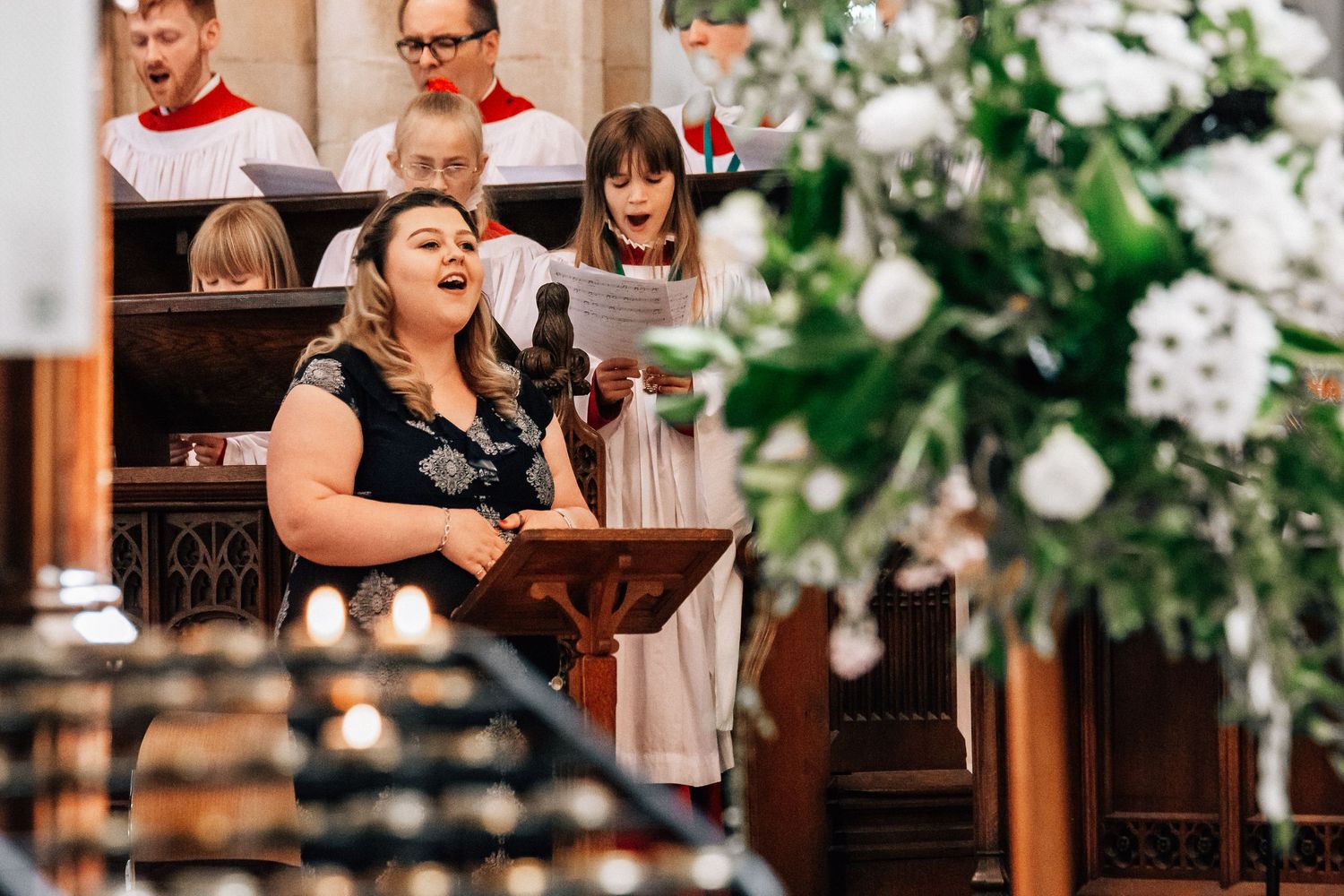 A woman sings joyfully at a church wedding ceremony while choir members perform in white robes behind her.