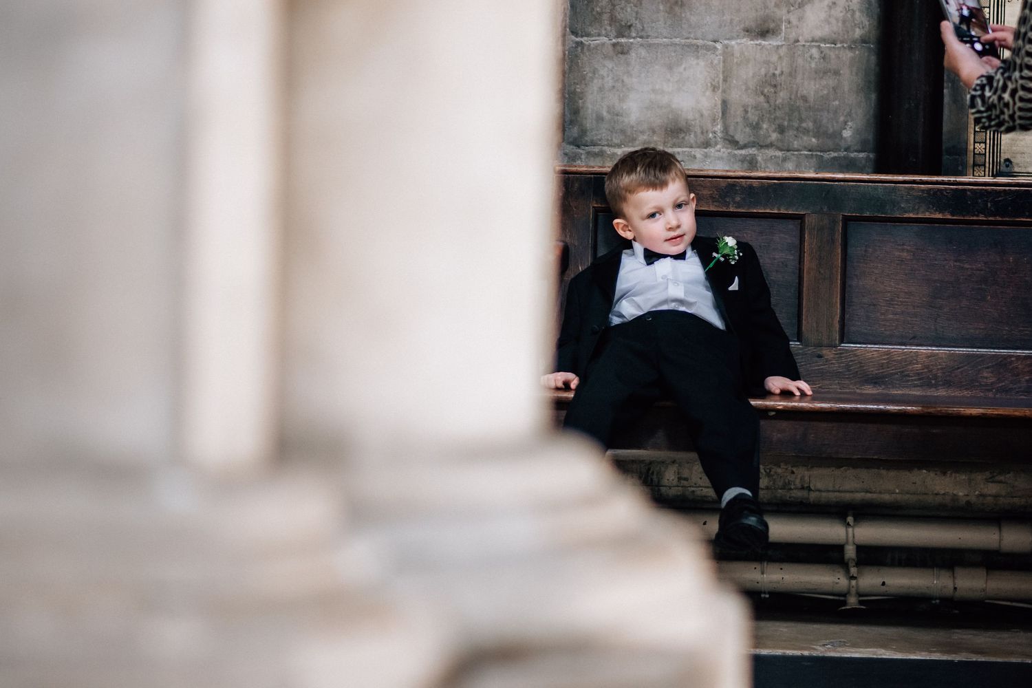 A young boy in a black tuxedo sits relaxed on a wooden church bench during a wedding ceremony.