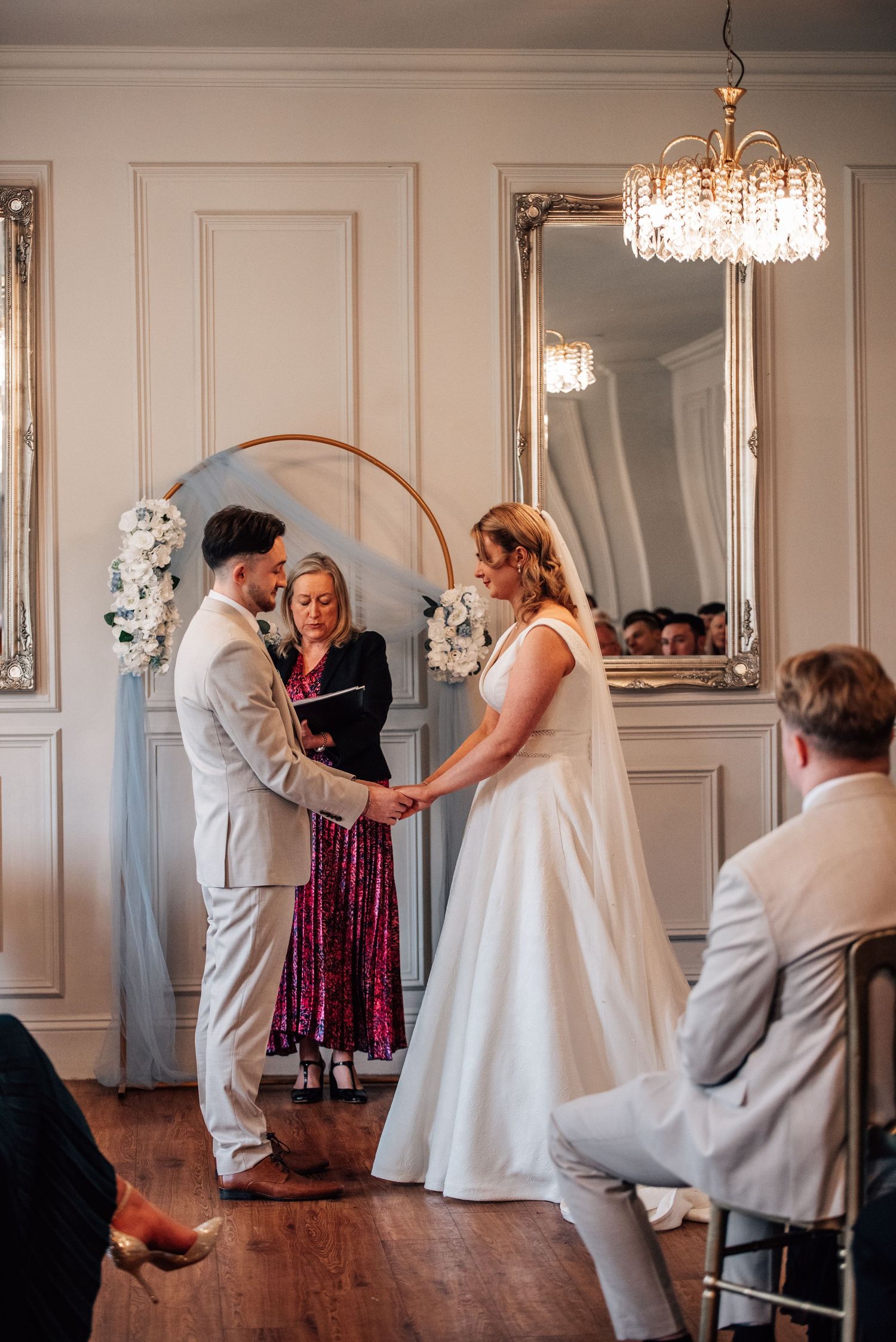 Couple exchanging vows at indoor wedding ceremony with floral arch, officiant, and elegant mirrored walls.