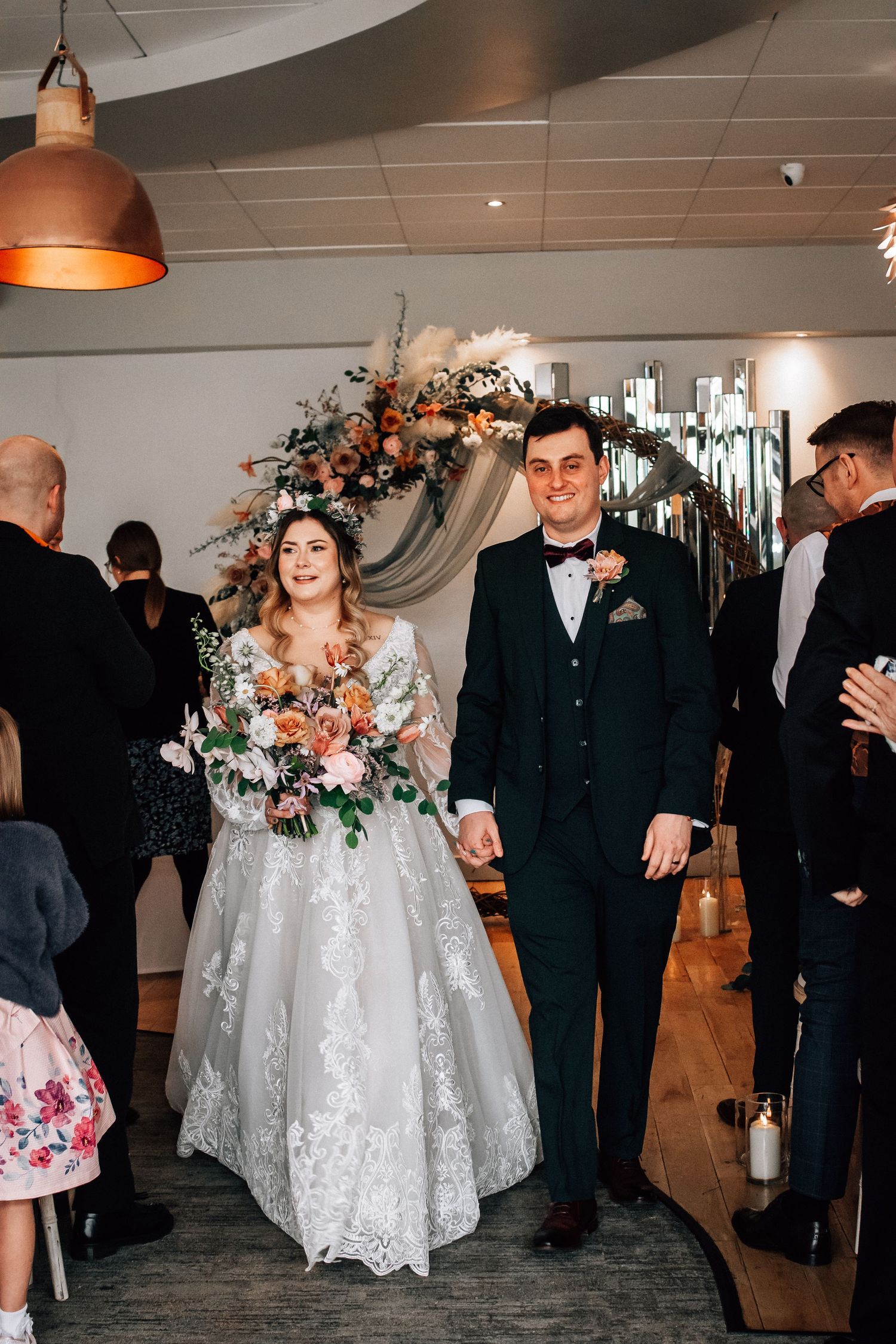 Bride and groom walk down the aisle smiling, holding hands, with floral arch and wedding guests in background.