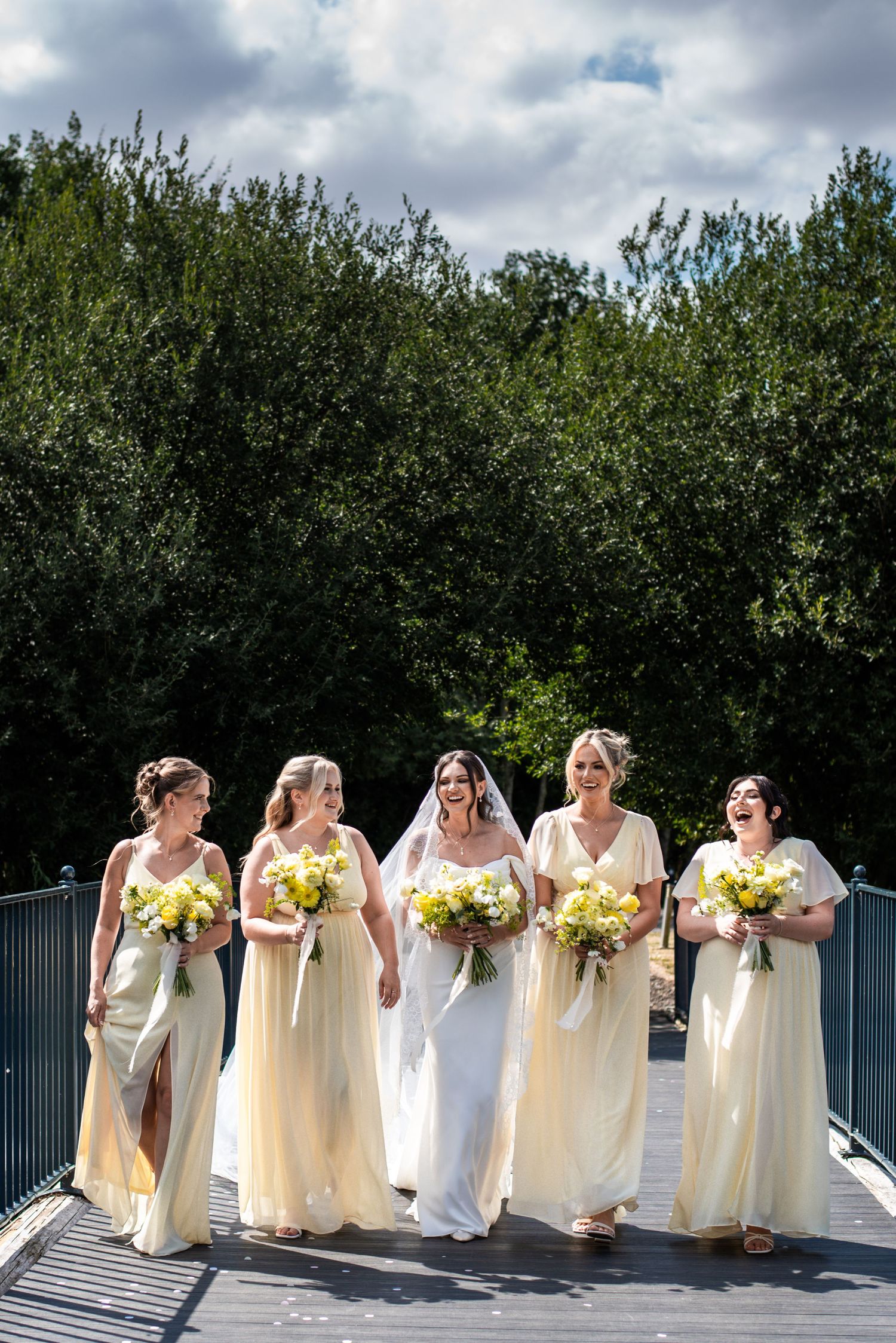 Bride in white gown walks with four bridesmaids in yellow dresses carrying bouquets on bridge.