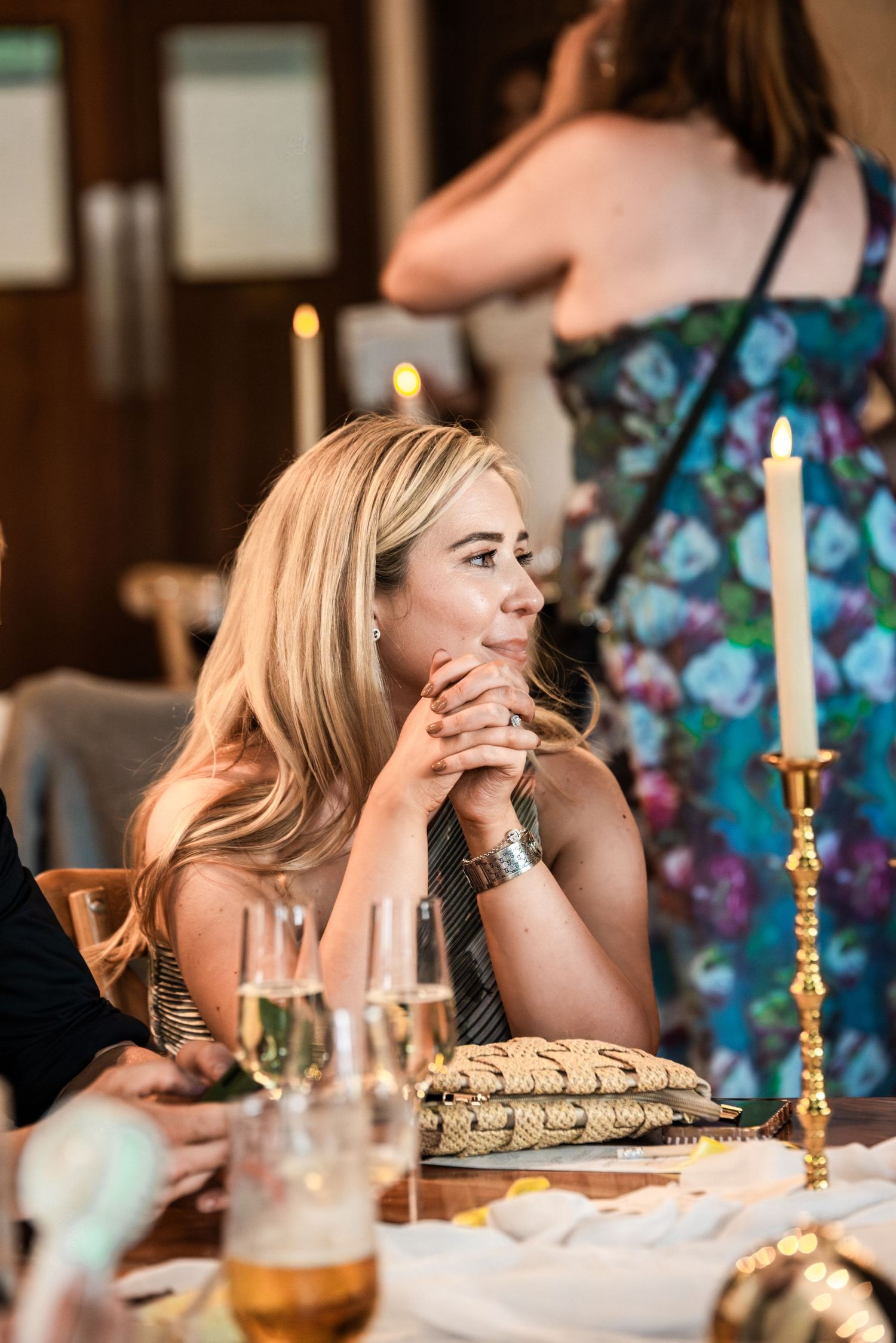 Blonde woman in elegant attire sits at a candlelit dinner table, hands clasped, listening attentively at a formal event.