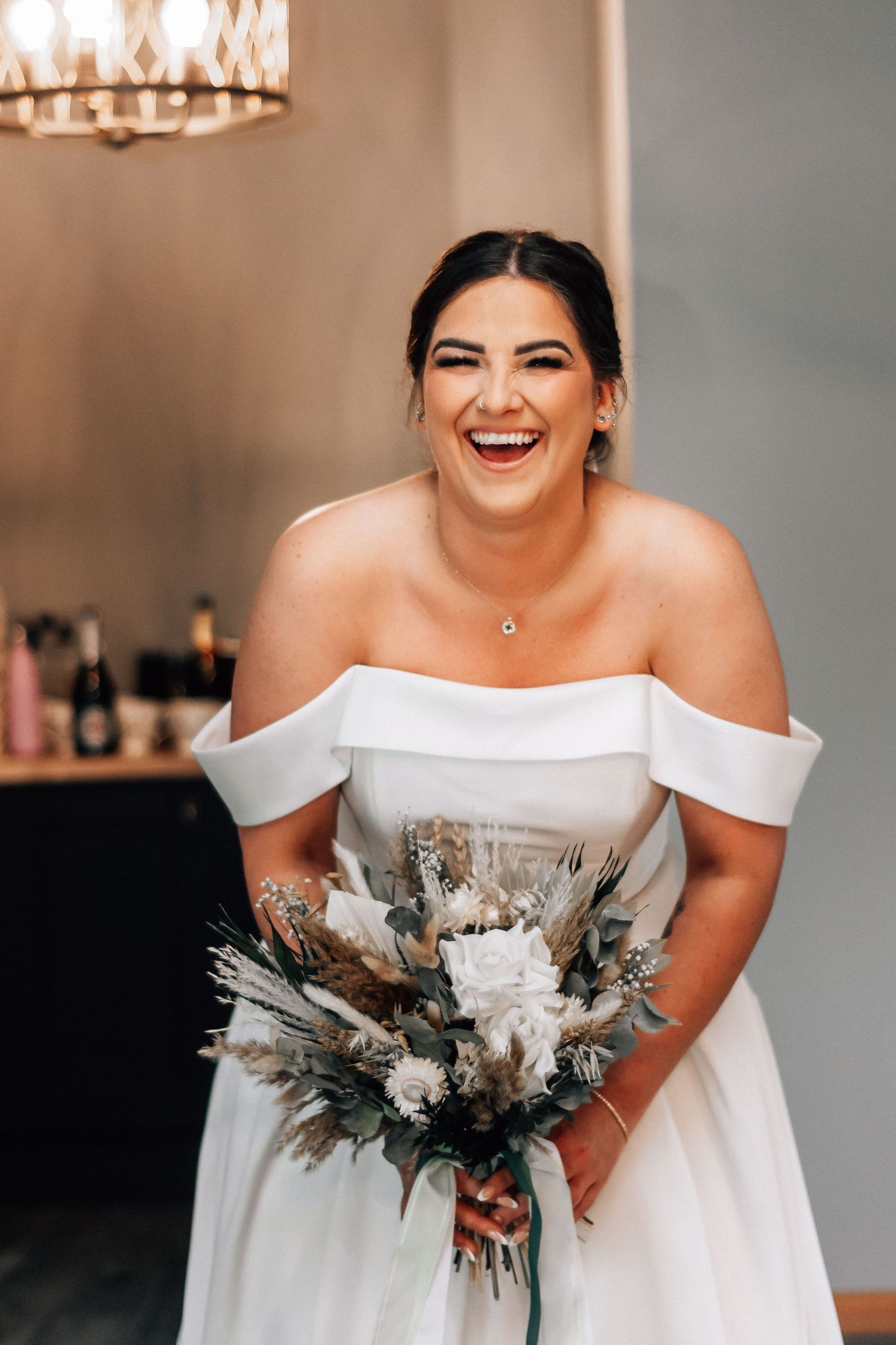 Laughing bride in off-shoulder white gown holds boho bouquet of dried flowers and greenery indoors.
