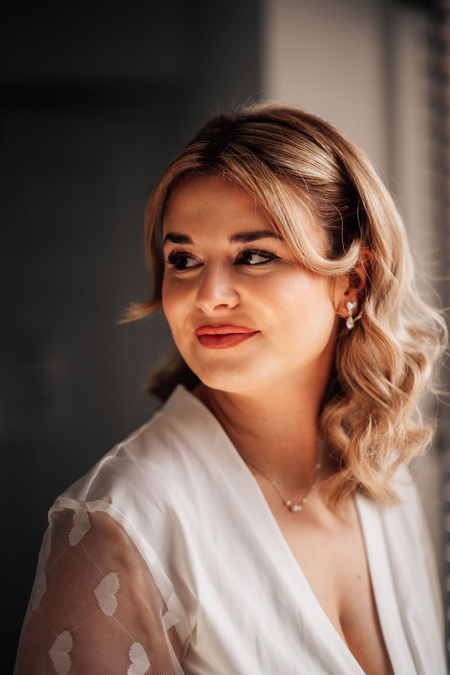 Elegant bride with blonde hair, red lips, pearl earrings, and necklace wearing a white robe before her wedding.