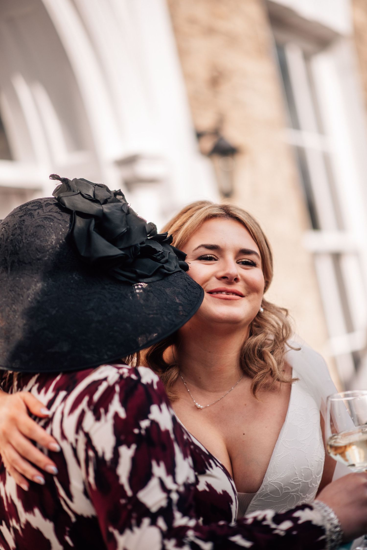 A smiling bride in white holds champagne while being kissed on the cheek by a guest in a black fascinator hat.
