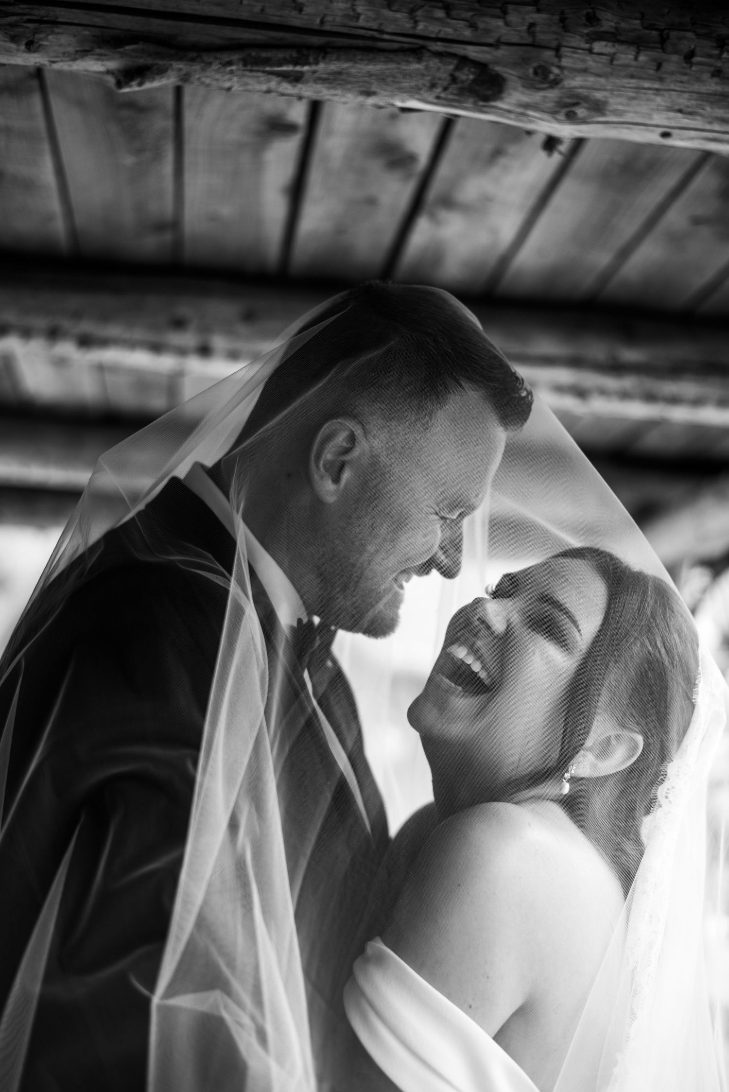 Black and white photo of a laughing bride and groom sharing a tender moment under her veil.