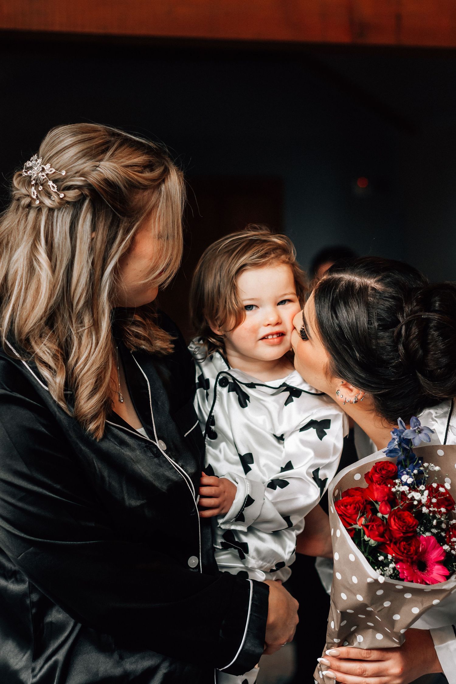 Bride in white robe kisses a smiling toddler held by a bridesmaid during wedding morning preparations.