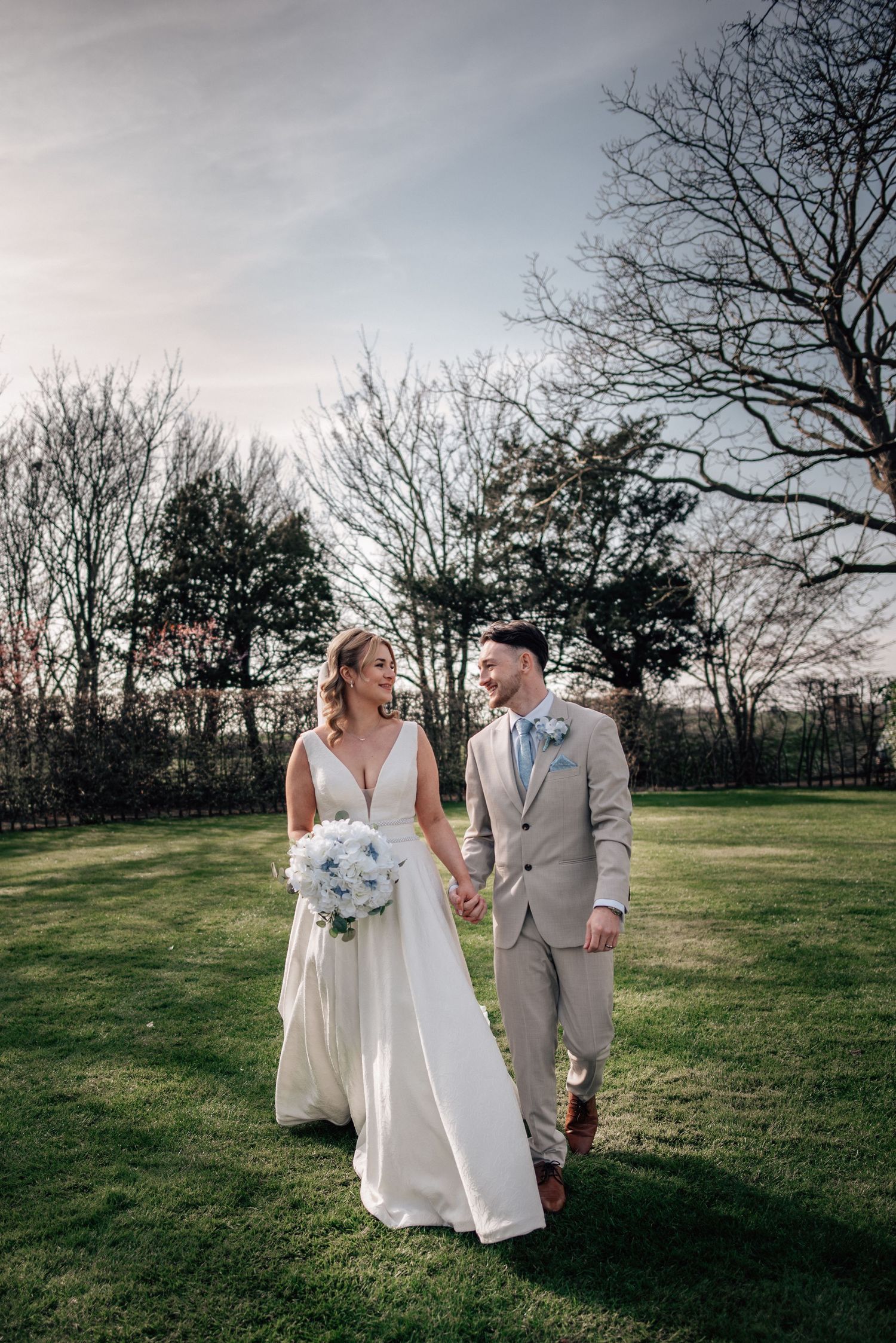 A bride and groom walk hand-in-hand on green grass, she in a white gown with bouquet, he in a tan suit.