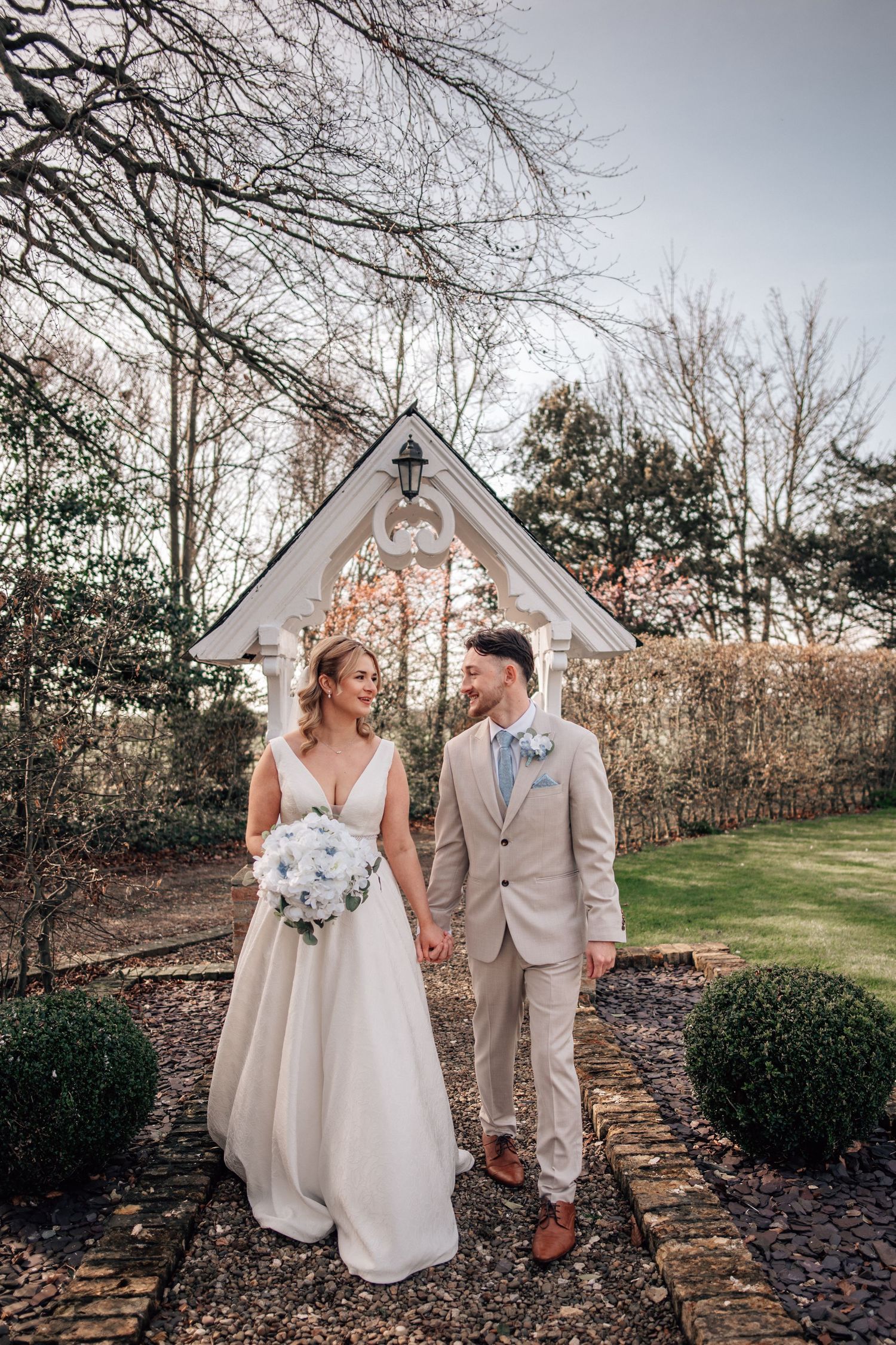 Bride and groom holding hands in a garden, bride in white gown with bouquet, groom in beige suit with blue bow tie.