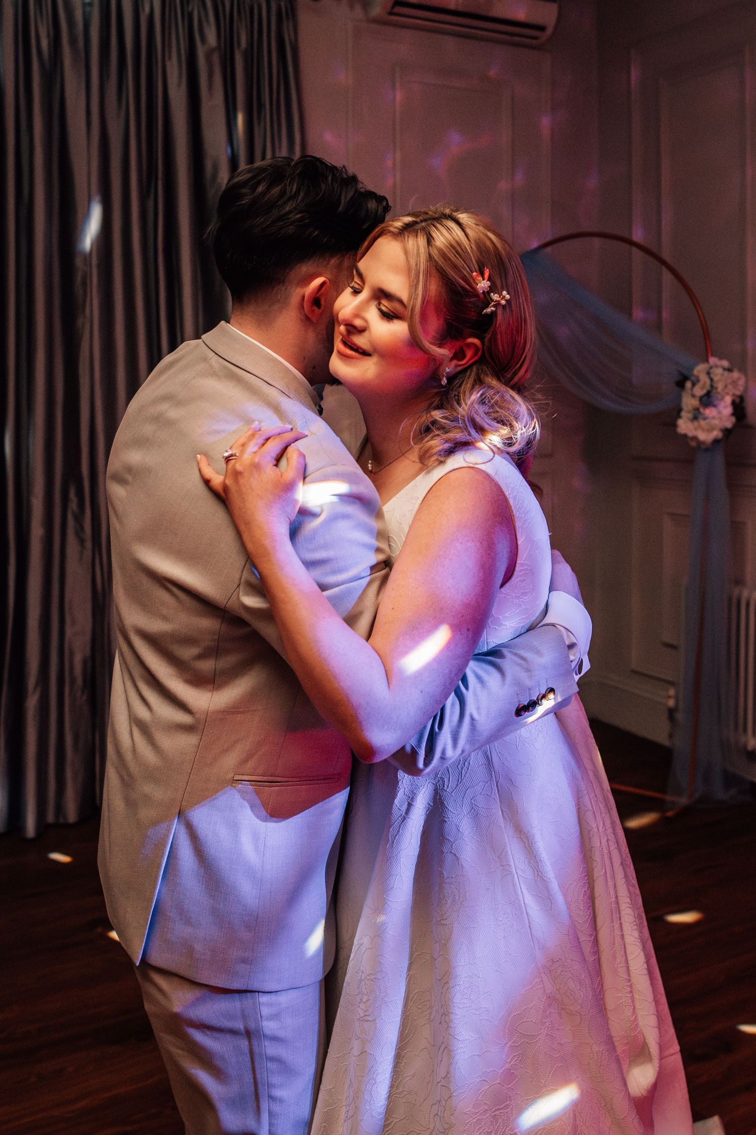 Newlywed couple shares an intimate first dance embrace in a softly lit wedding reception venue.