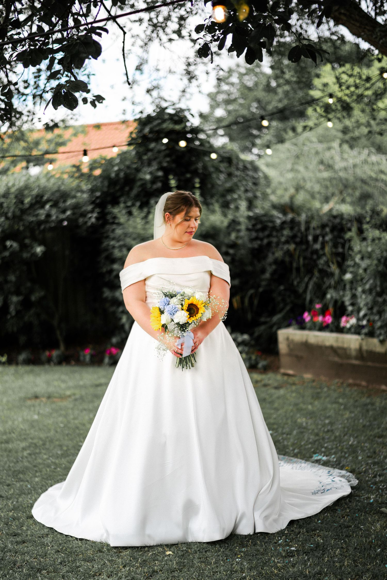 Bride in off-shoulder white ball gown holding sunflower bouquet stands in lush garden setting.