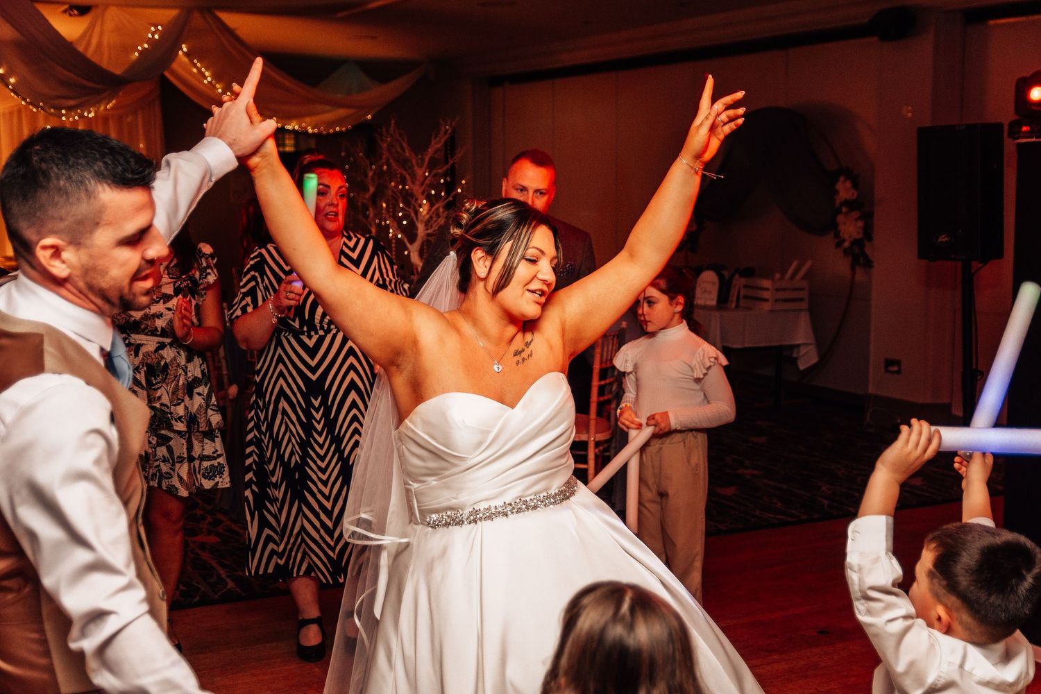 Bride in strapless wedding gown raises arms joyfully on dance floor surrounded by guests at lively wedding reception.