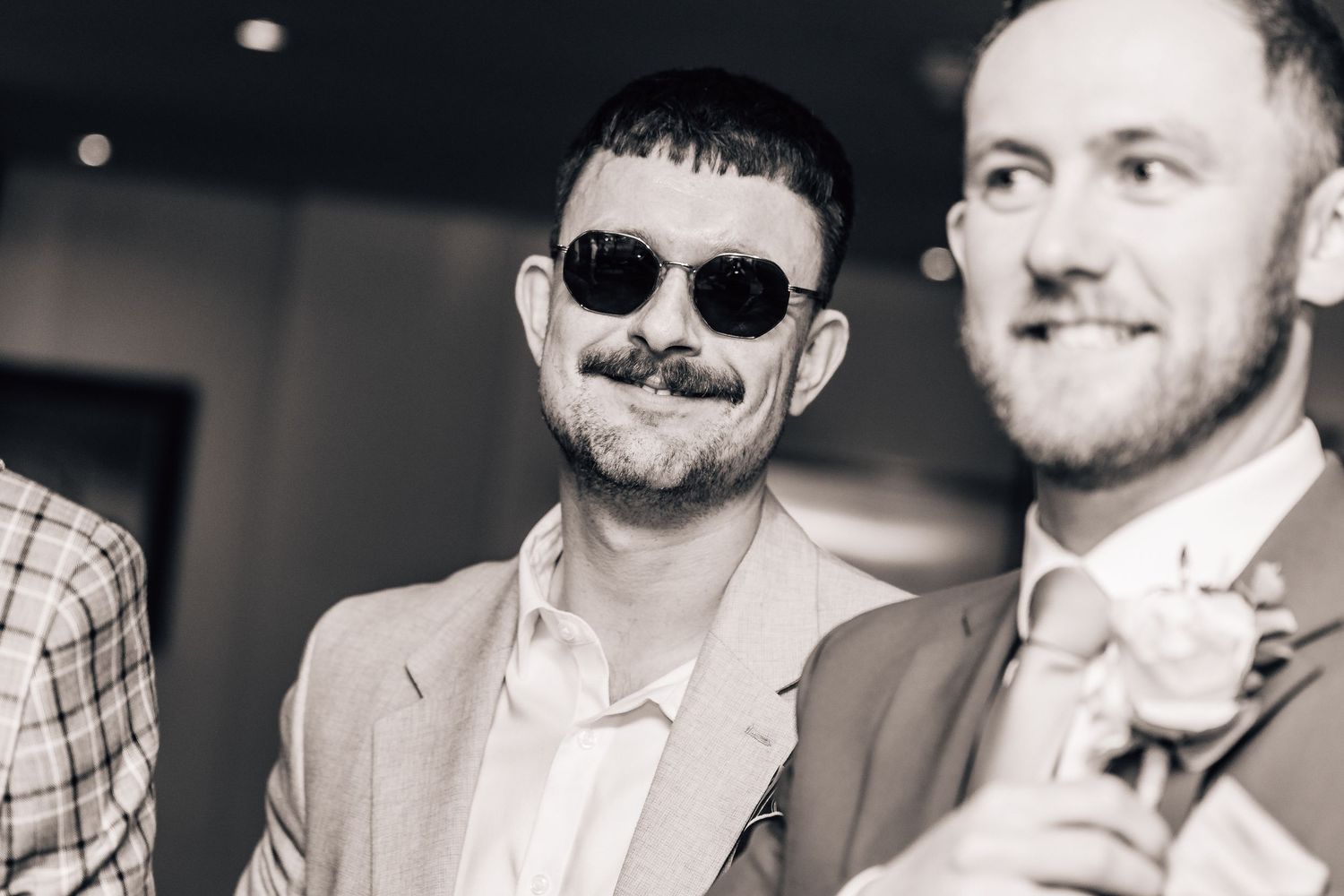 Black and white photo of two smiling men in suits at a wedding, one wearing round sunglasses.