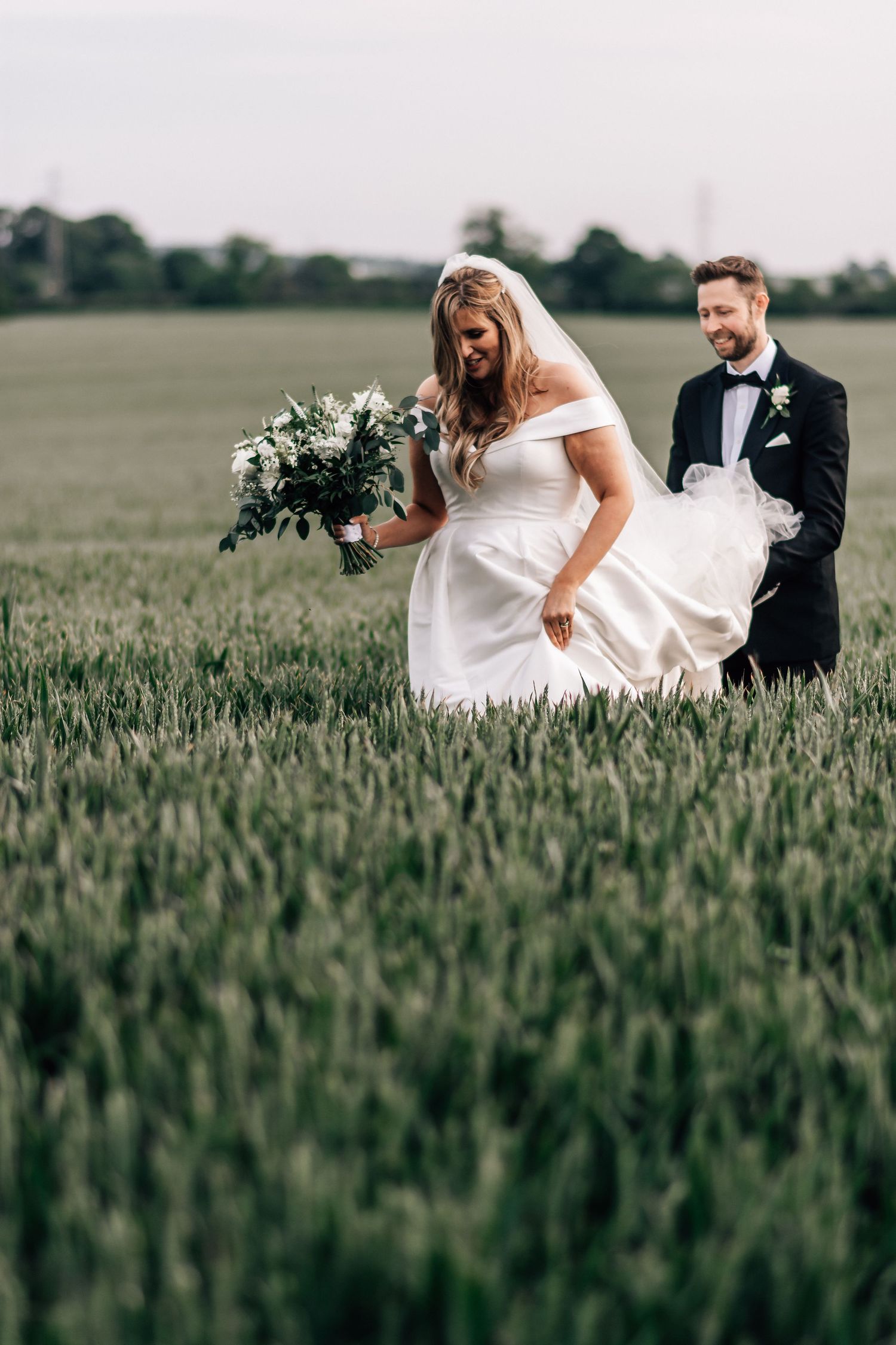 Bride and groom walking through a lush green field, bride holding a white floral bouquet in a flowing off-shoulder wedding dress.