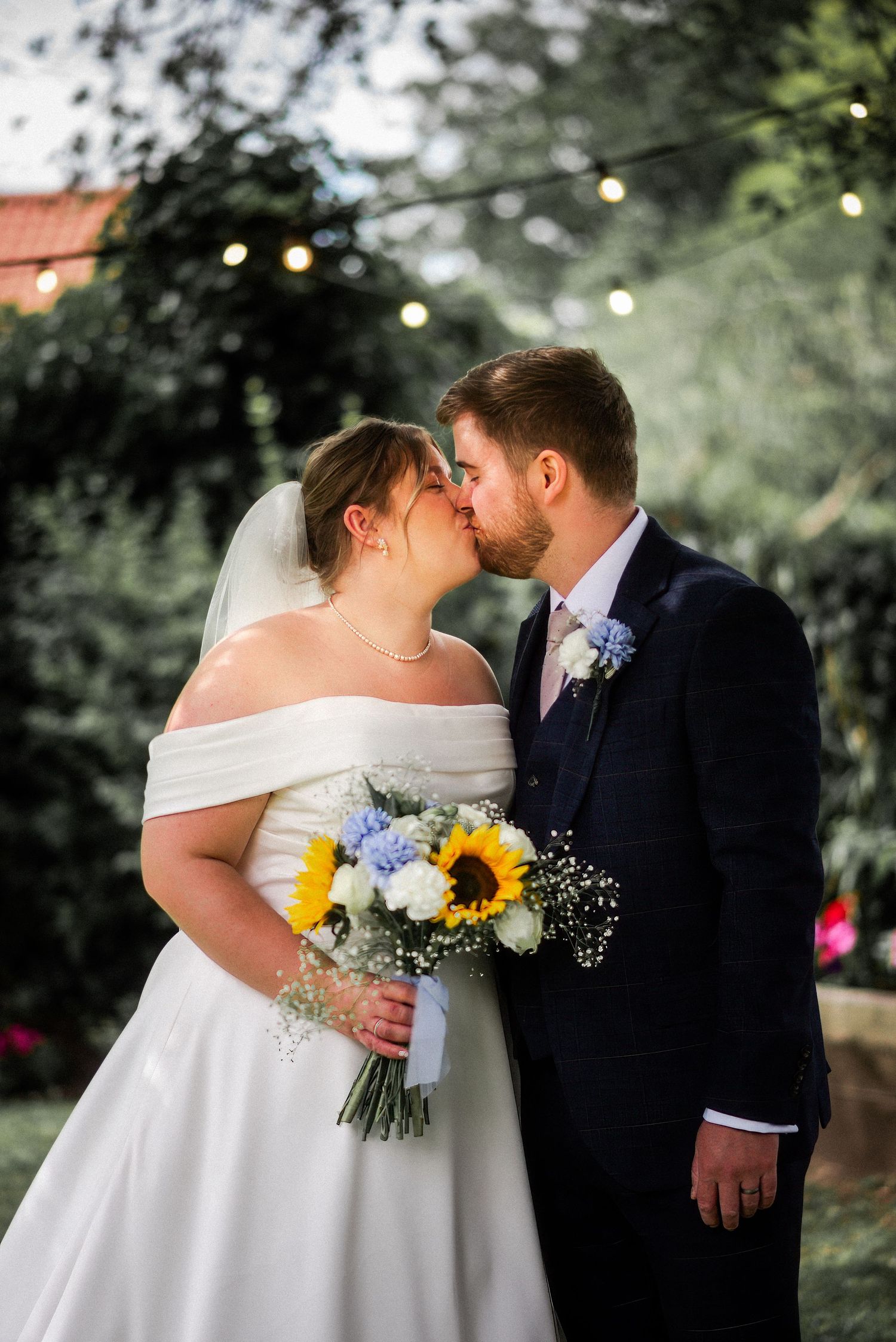 Bride and groom share a kiss outdoors, bride holding sunflower and blue hydrangea bouquet, groom in navy suit.
