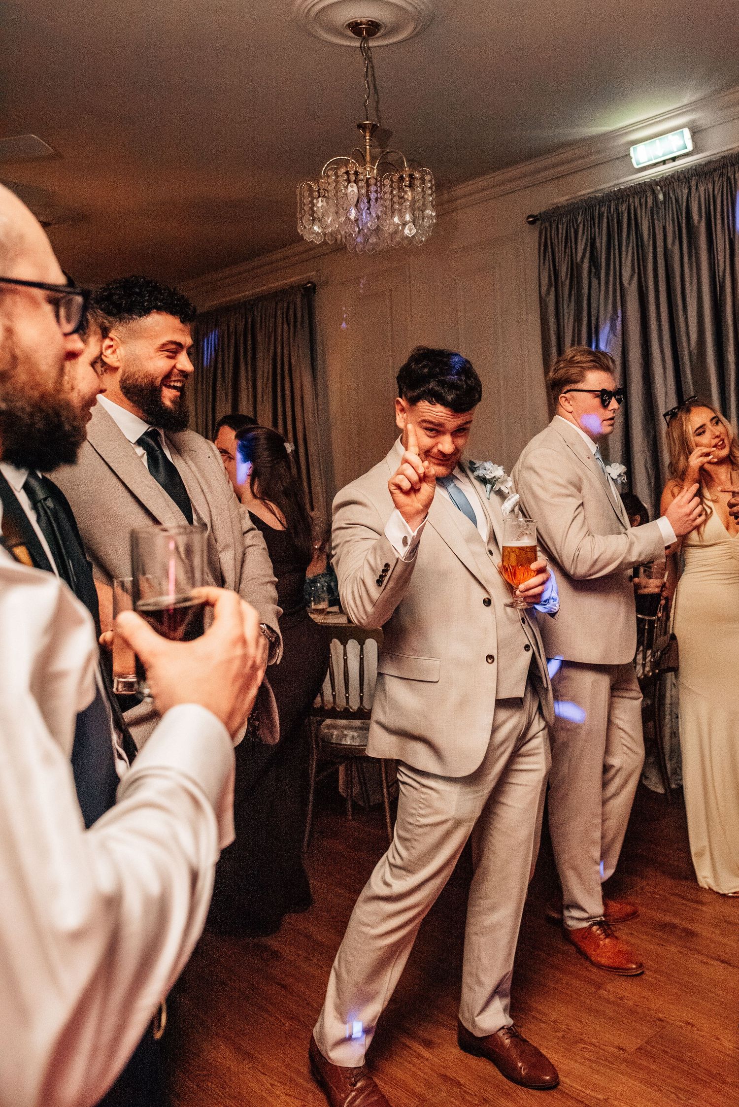 Groom in grey suit dancing joyfully with groomsmen and guests at lively wedding reception party.