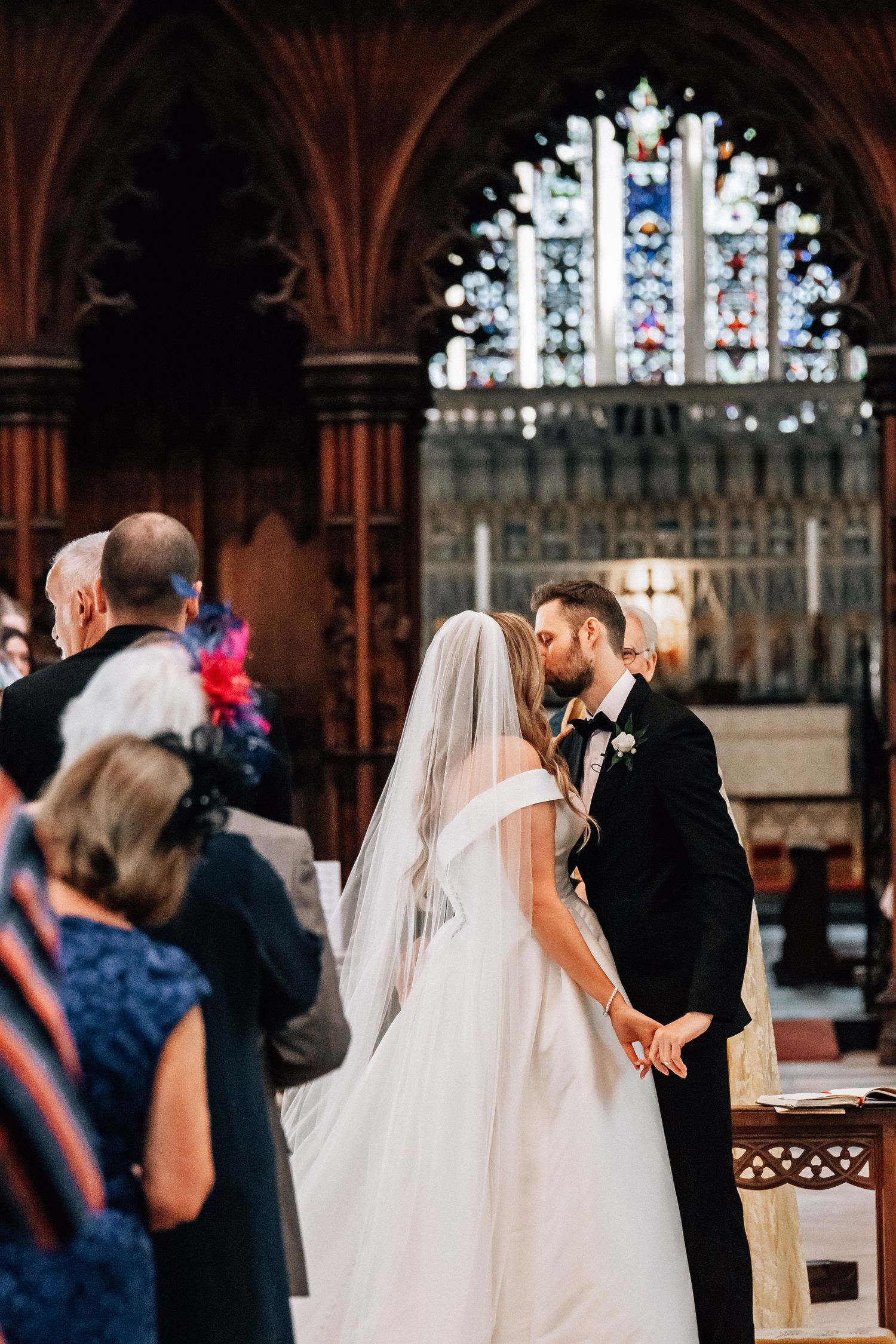 Bride and groom share first kiss at church altar with stunning stained glass window backdrop during wedding ceremony.