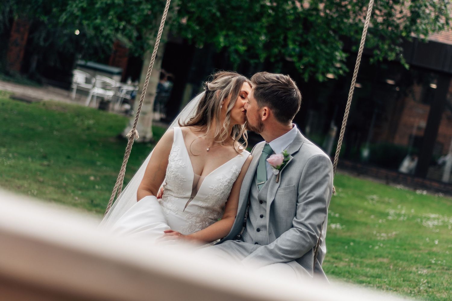 Bride and groom sharing a romantic kiss on a wooden swing in a lush green garden on their wedding day.