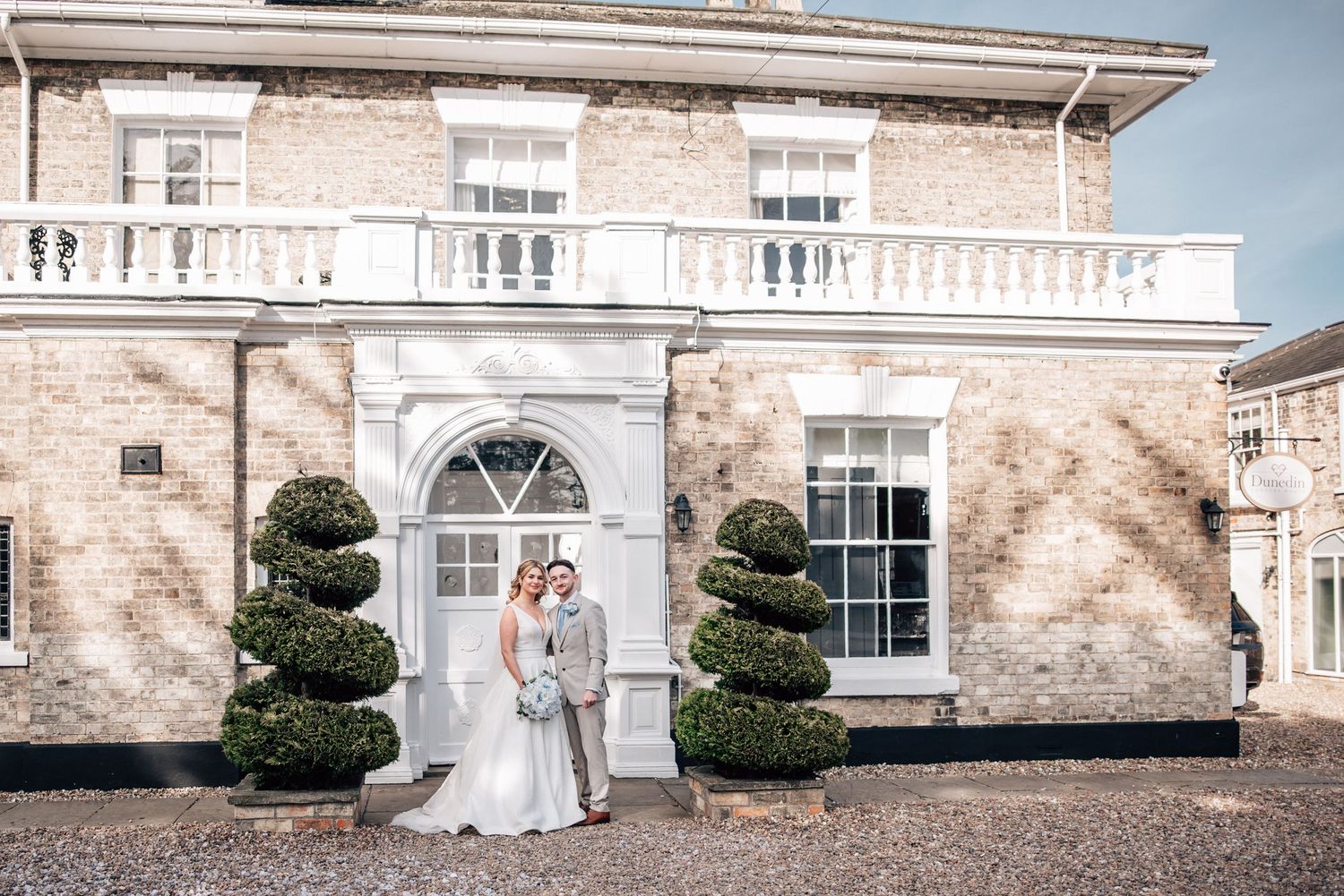 Bride and groom pose together outside an elegant brick manor house with white columns and topiary bushes.