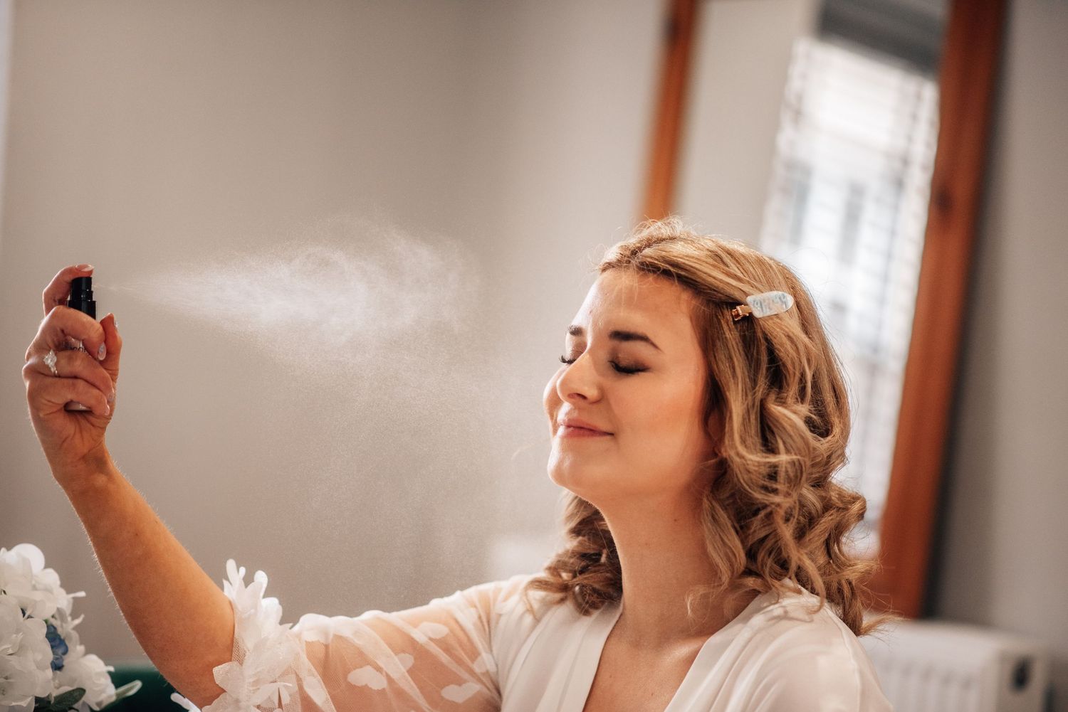 Bride in white robe spraying facial mist with eyes closed, preparing for wedding day beauty routine.