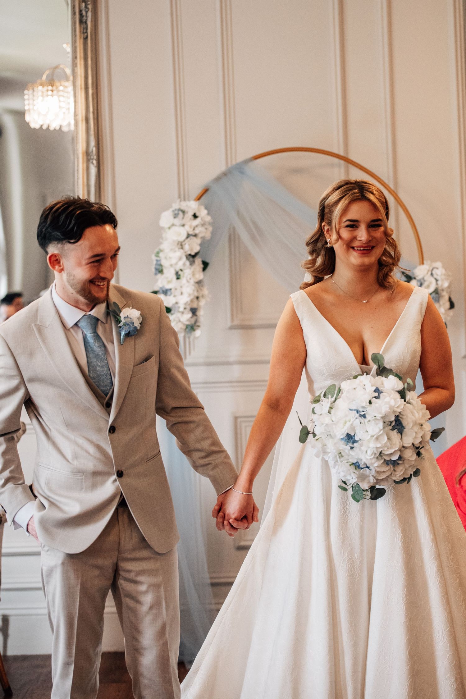 Smiling bride in white gown holding bouquet walks hand-in-hand with groom in beige suit at elegant wedding ceremony.