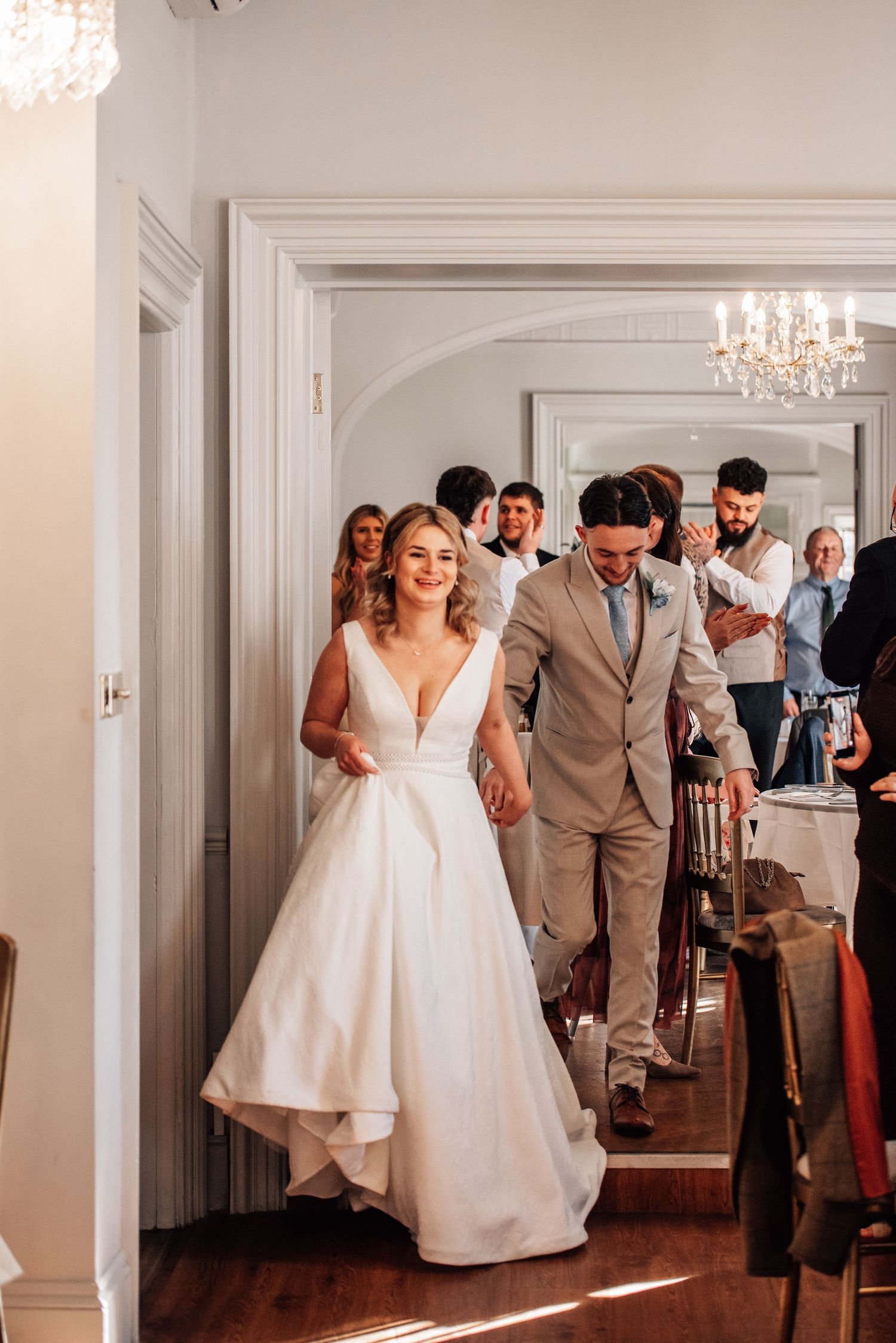 Bride and groom make grand entrance at wedding reception, guests applauding in elegant white hallway.