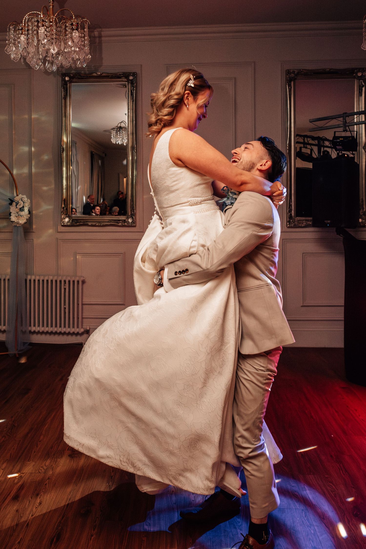 Groom lifting laughing bride in white wedding dress during energetic first dance at reception.