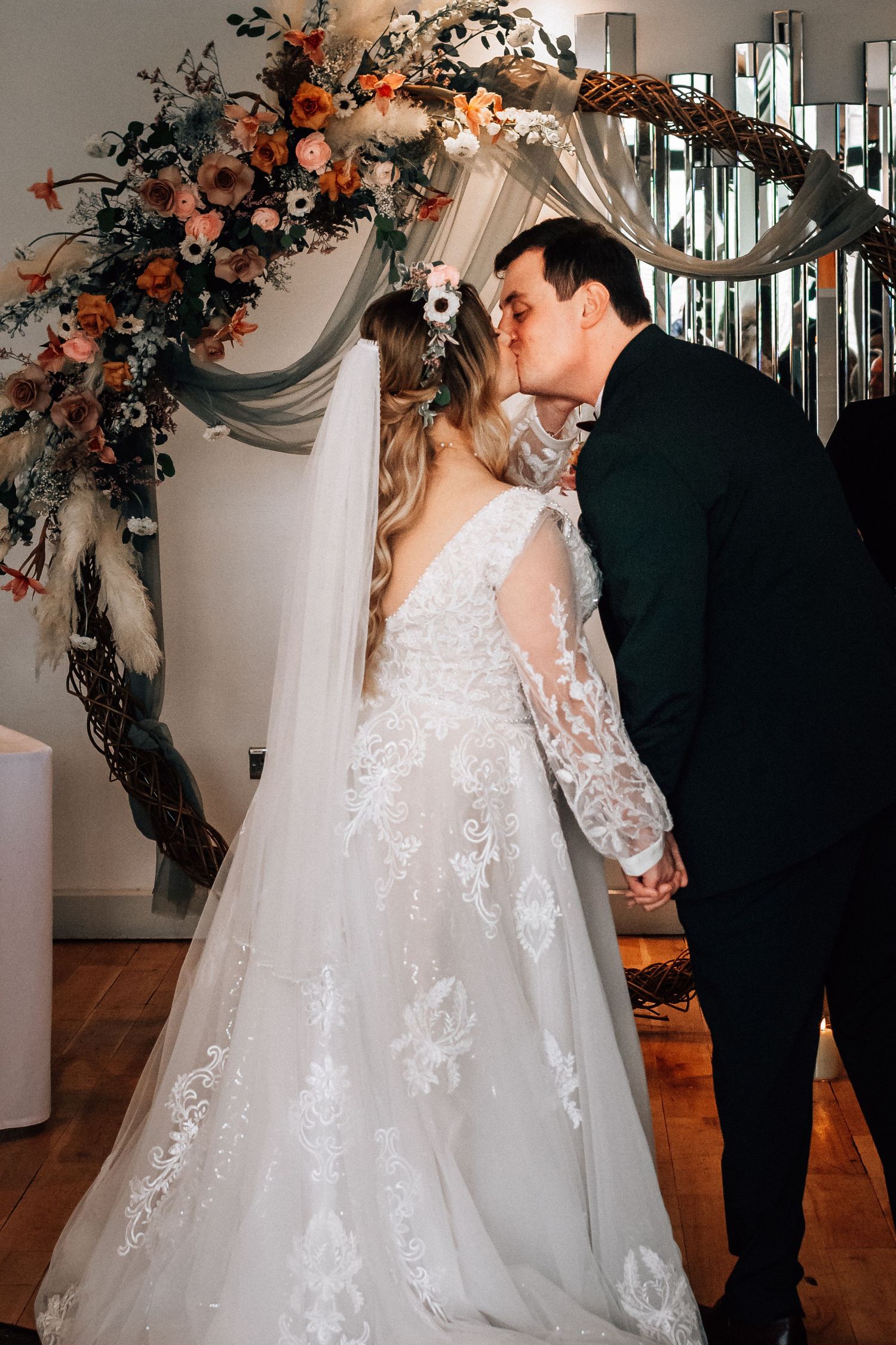 Bride and groom share first kiss at wedding altar adorned with boho floral arch in orange and white blooms.