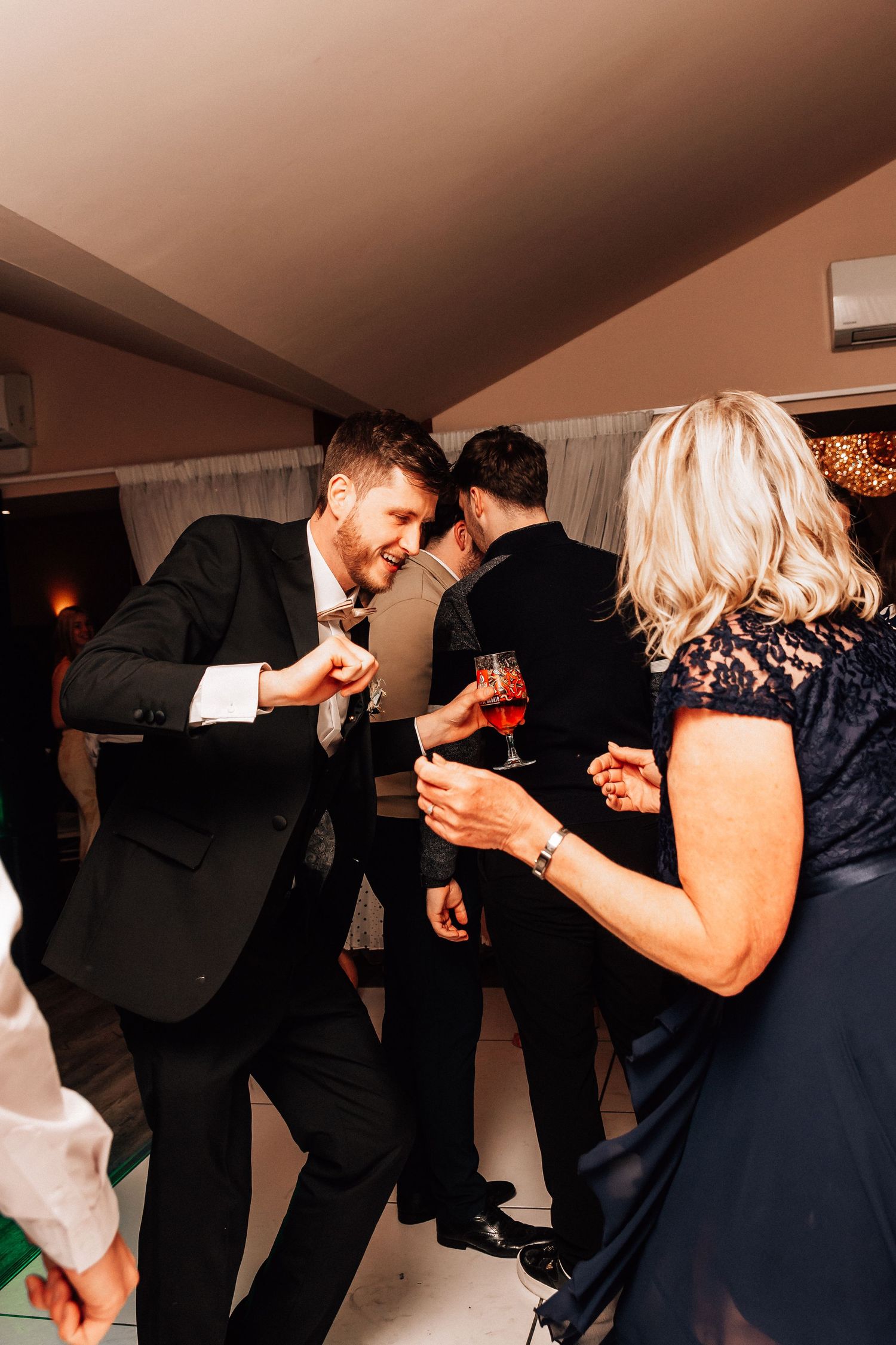 Smiling wedding guest in black suit dances energetically with guests at a lively wedding reception.