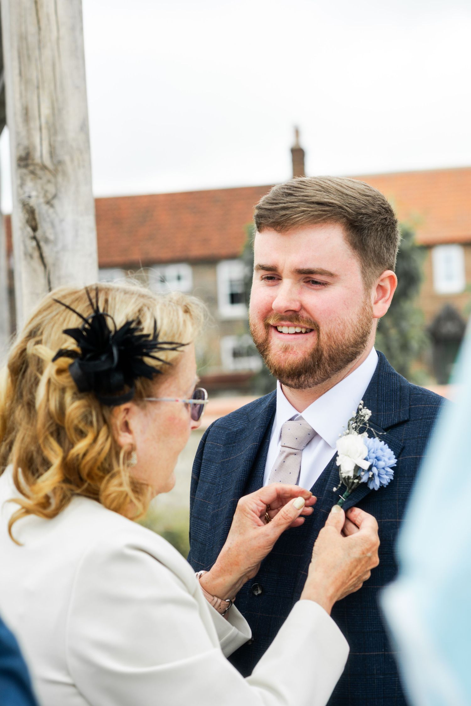 A woman pins a boutonniere on a smiling bearded groom in a navy suit before a wedding ceremony.