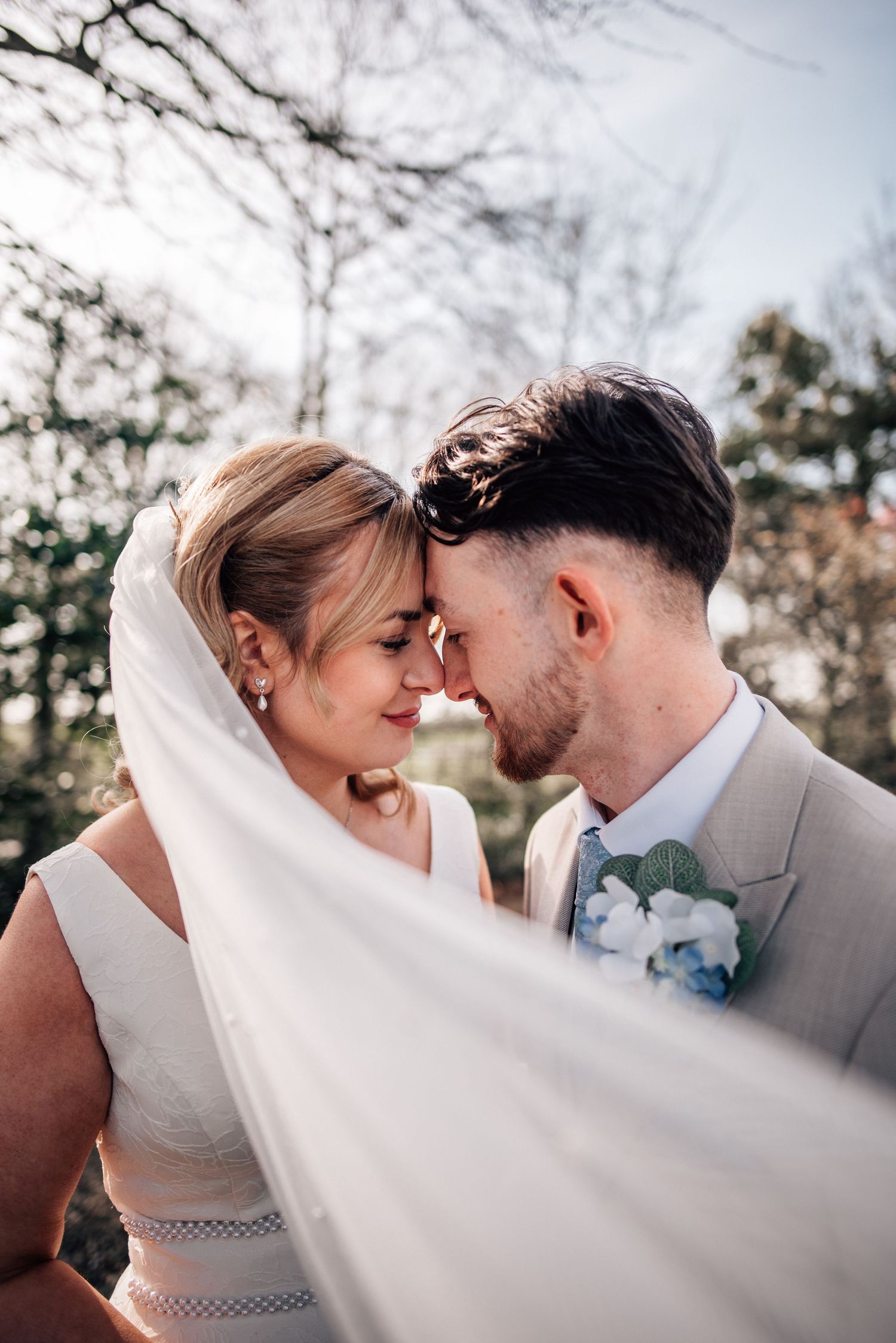 A bride and groom touch foreheads tenderly outdoors, with a flowing white veil sweeping across the foreground.