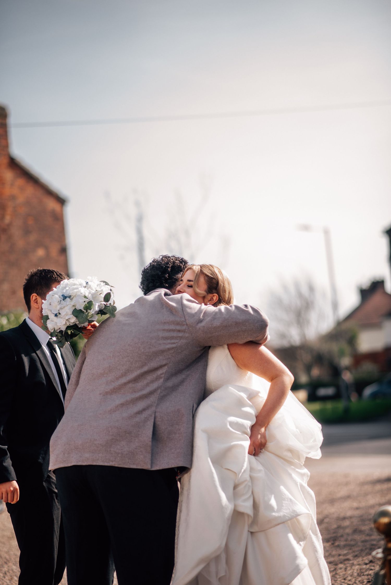 Bride in white gown hugging a guest in gray suit outdoors, with another man in black suit holding a bouquet nearby.